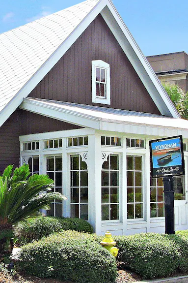 A house with a steep roof and a small upper window, featuring a glass-enclosed porch with white framing, surrounded by bushes and a black real estate sign at Wyndham Vacation Rentals.