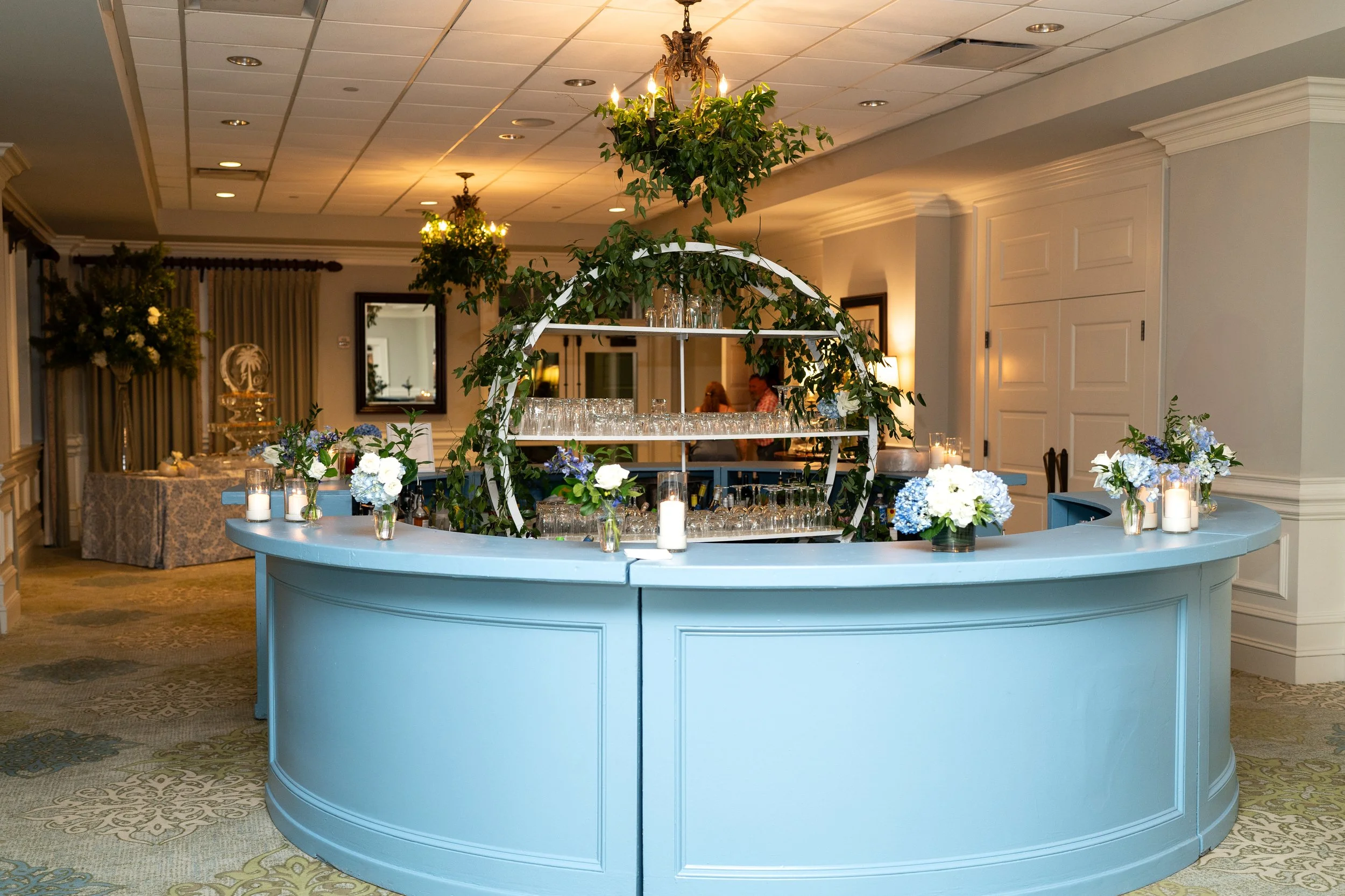 Pictured in the Carolina Room at Seabrook Island Club is an elegant blue bar decorated with white and blue flowers, candles, and glassware, with hanging chandeliers and floral arrangements in the background.