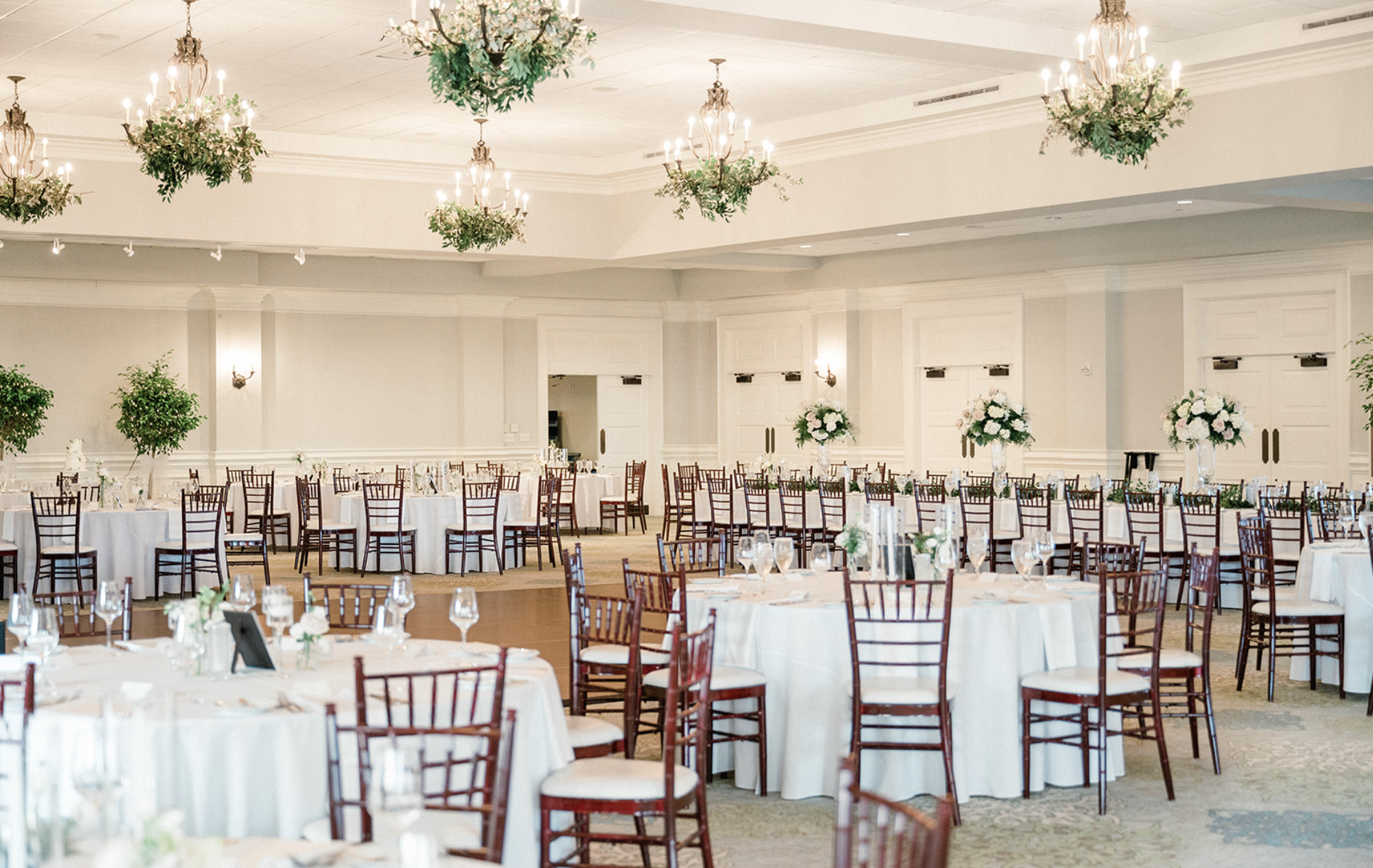 The Atlantic Ballroom at Seabrook Island Club elegantly decorated for a wedding reception with round tables draped in white tablecloths, surrounded by wooden chairs, and decorated with tall floral centerpieces and chandeliers.