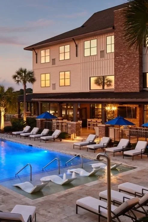 A luxurious outdoor pool area at sunset with lounge chairs, umbrellas, and a large two-story building in the background featuring many windows and palm trees at the Andell Inn.