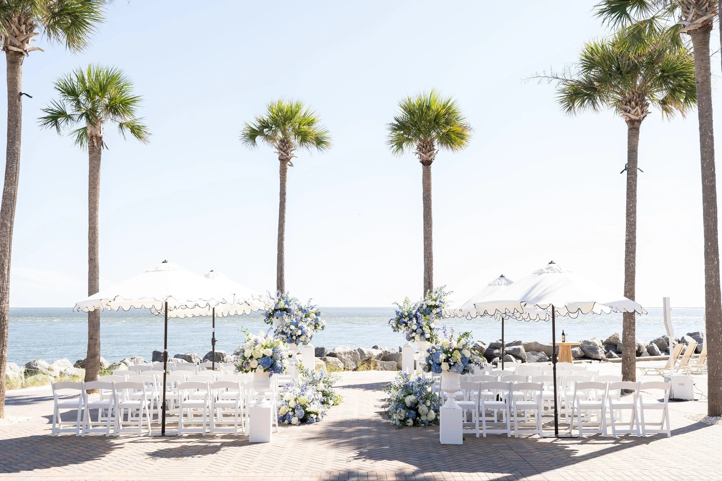 Beachside wedding setup on Seabrook Island Club's Ocean Terrace with white chairs, floral arrangements, umbrellas, palm trees, and ocean in the background.