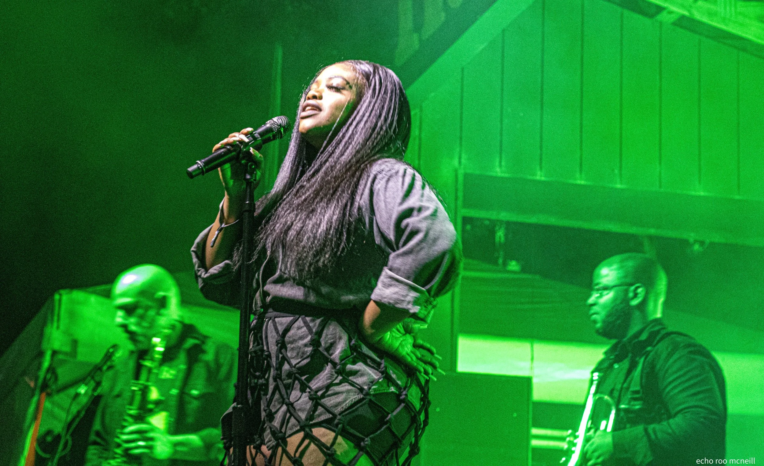A woman singing into a microphone on stage, with green lighting and two musicians playing instruments in the background.