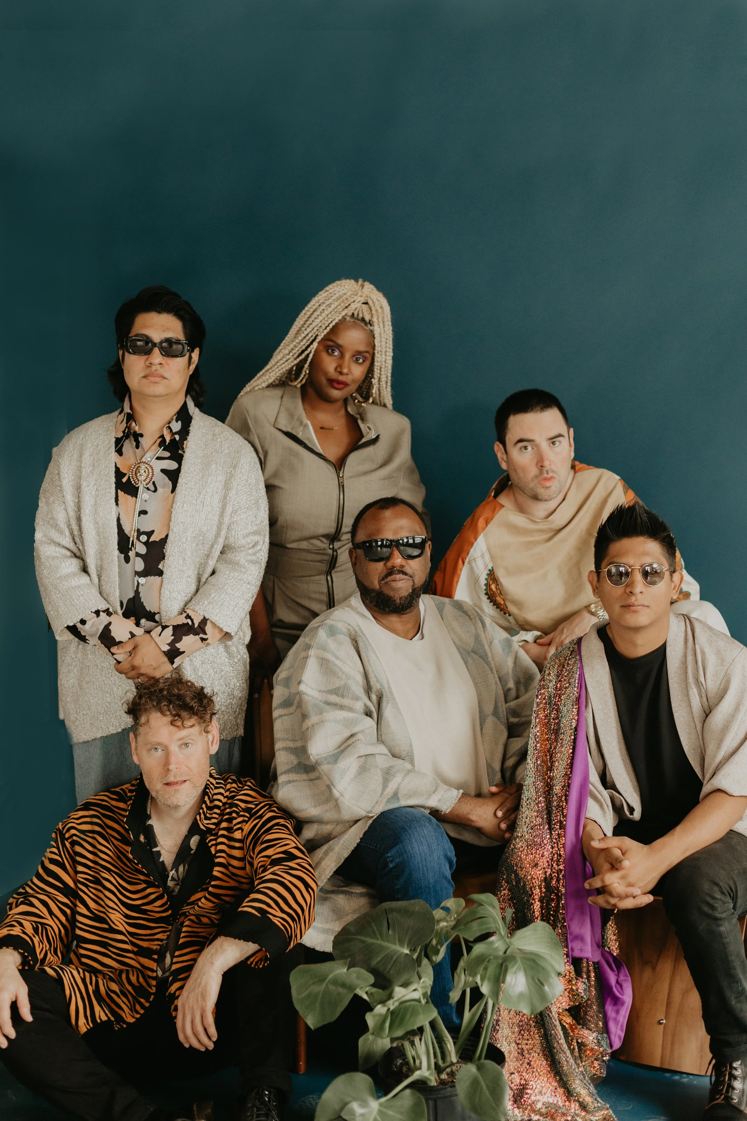 Group of seven diverse individuals posing together indoors against a blue background, some wearing sunglasses, casual and eclectic fashion styles.
