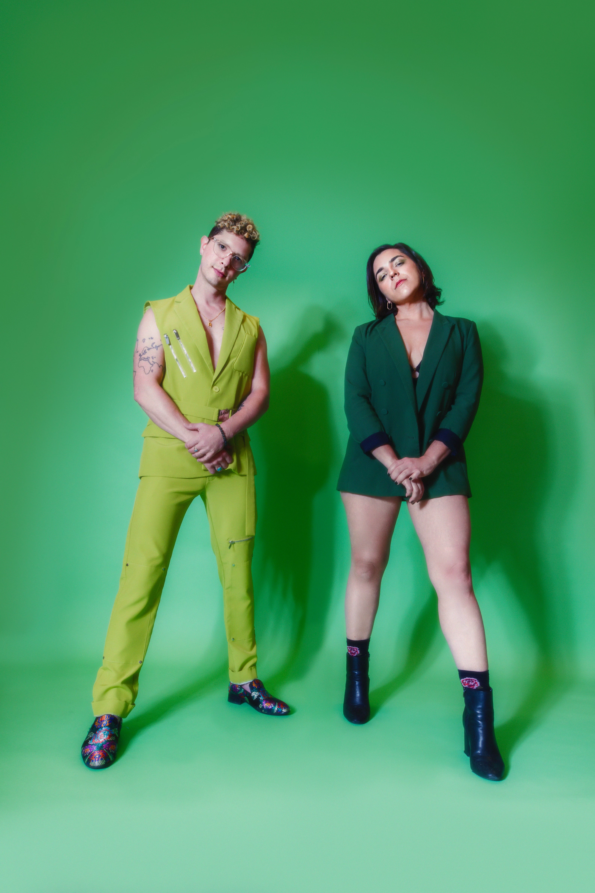 Two individuals standing against a green screen background, wearing colorful and stylish outfits. The person on the left has short curly hair, glasses, tattoos, and is dressed in a lime green sleeveless vest and matching pants with floral shoes. The person on the right has shoulder-length black hair, and is dressed in a dark green blazer with no visible shirt or pants, and is wearing black leather ankle boots and patterned socks.