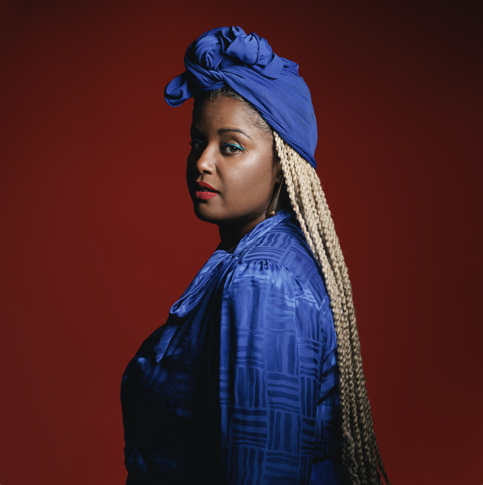 Portrait of a woman with long blonde braids wearing a blue headwrap and a matching blue patterned blouse, with a red background.