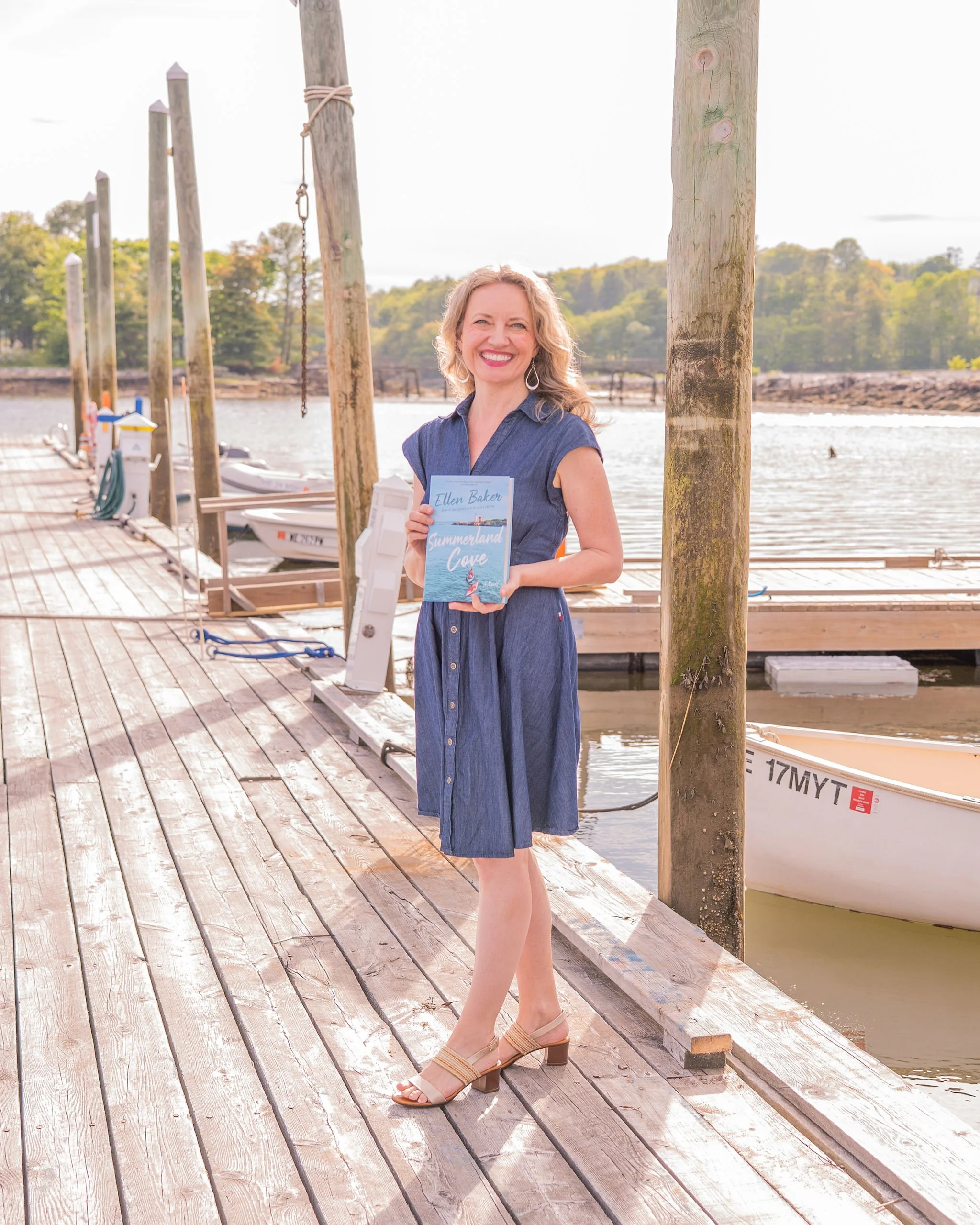 Ellen Baker in a navy dress holding a copy of Summerland Cove on a wooden dock, with boats and the harbor behind her.