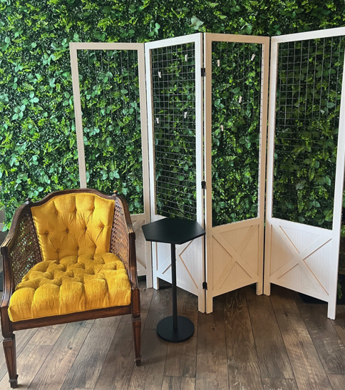 A yellow upholstered armchair with wooden frame, a small black side table, and a white four-panel folding screen with grid patterns, set against a green leafy wall on a wood floor.