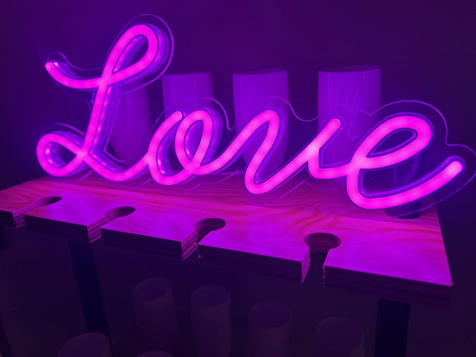 A neon sign spelling 'Love' in pink and purple colors, mounted on a dark wall with several cylindrical objects in the background.