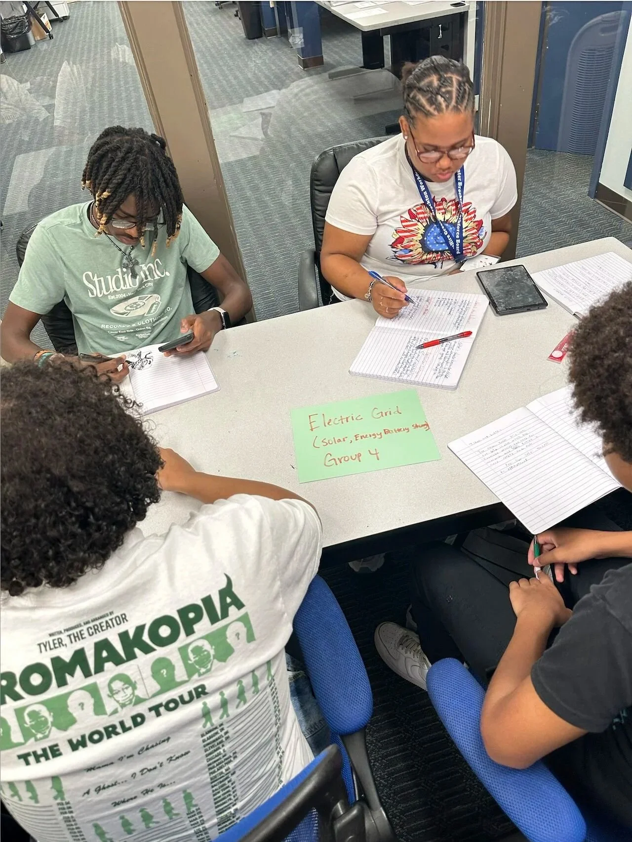 A group of students sitting around a table working on a project together. They are writing in notebooks, using a smartphone, and writing on a paper. The setting is a study area with glass walls and office furniture.
