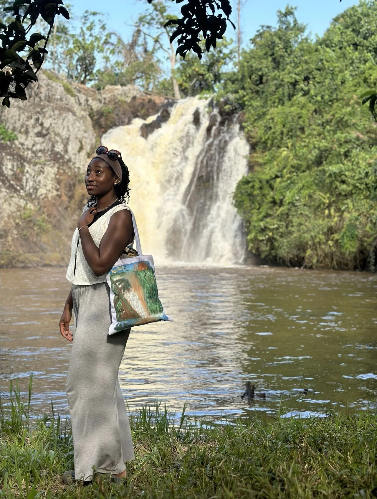 A woman standing on grass by a river with a waterfall in the background surrounded by green trees.