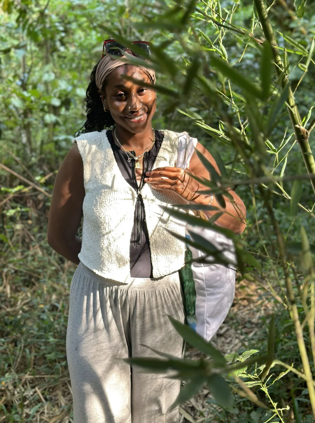 A woman smiling in a green, leafy outdoor setting, wearing a cream-colored sleeveless vest, gray pants, and sunglasses on her head.