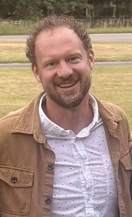 A smiling man with curly hair and a beard, wearing a white polka-dotted shirt and a brown jacket, standing outdoors on a grassy area with trees and a road in the background.