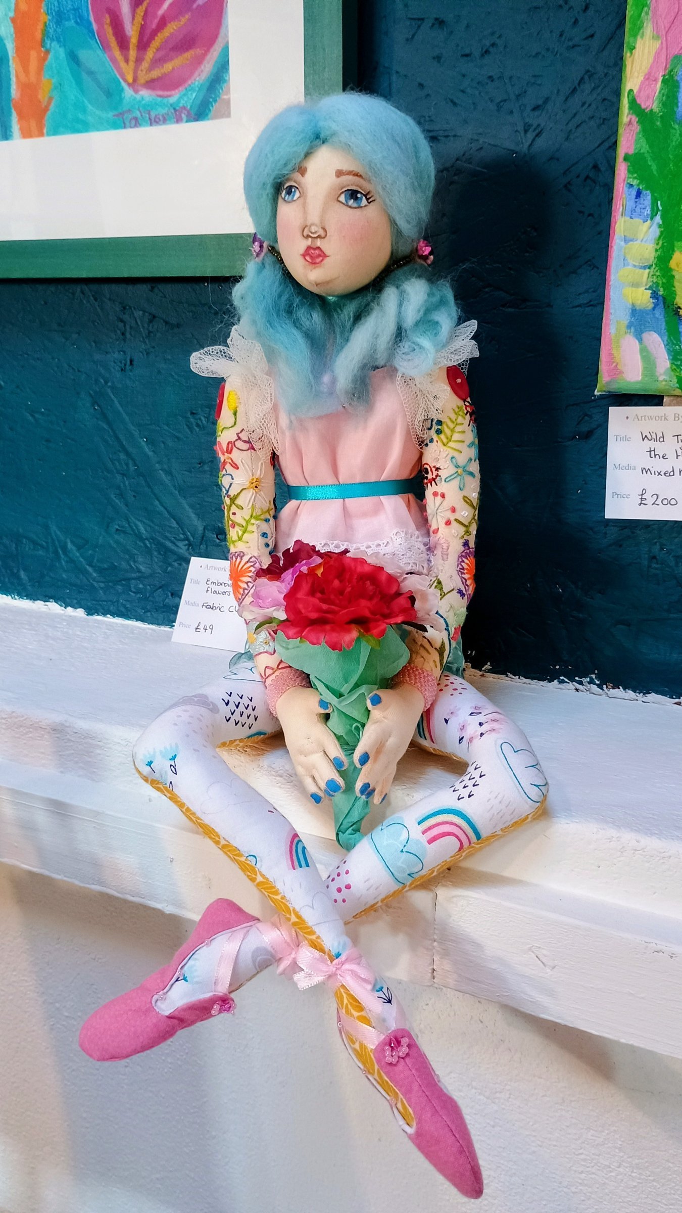 Handmade art doll with blue hair holding a red flower, sitting among Cerisia Ta'Torin paintings in the Ta'Torin Gallery, Hayle