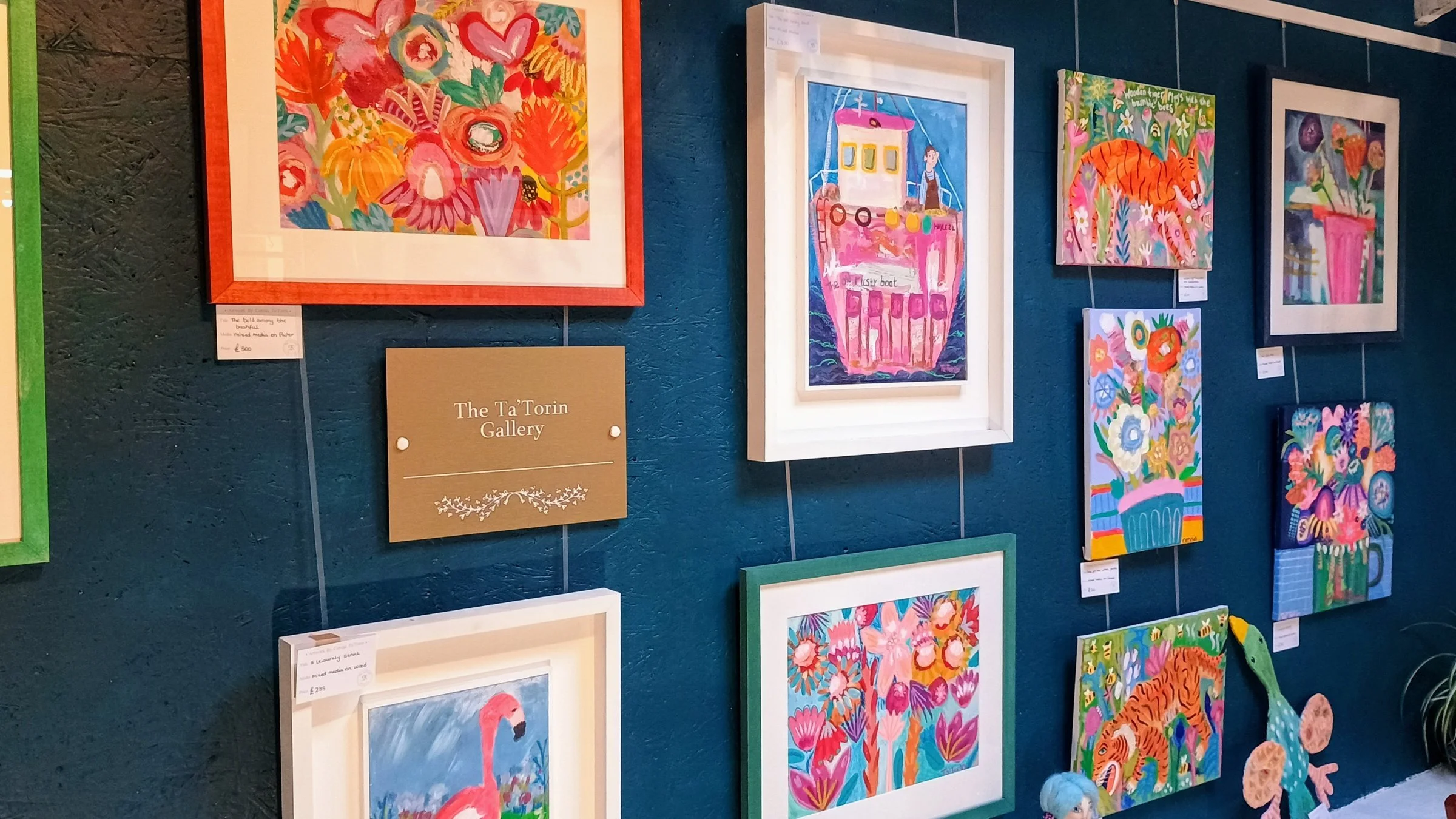 Colourful framed paintings by Cerisia Ta'Torin on a teal wall beside the Ta'Torin Gallery sign, inside Hayle Emporium