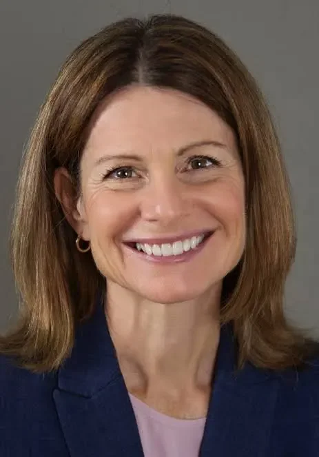 A photo of a woman with shoulder length light brown hair smiling and wearing a navy blue blazer with a lavender shirt underneath.
