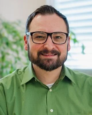 A smiling man is pictured wearing a green shirt with a plant and window behind him.