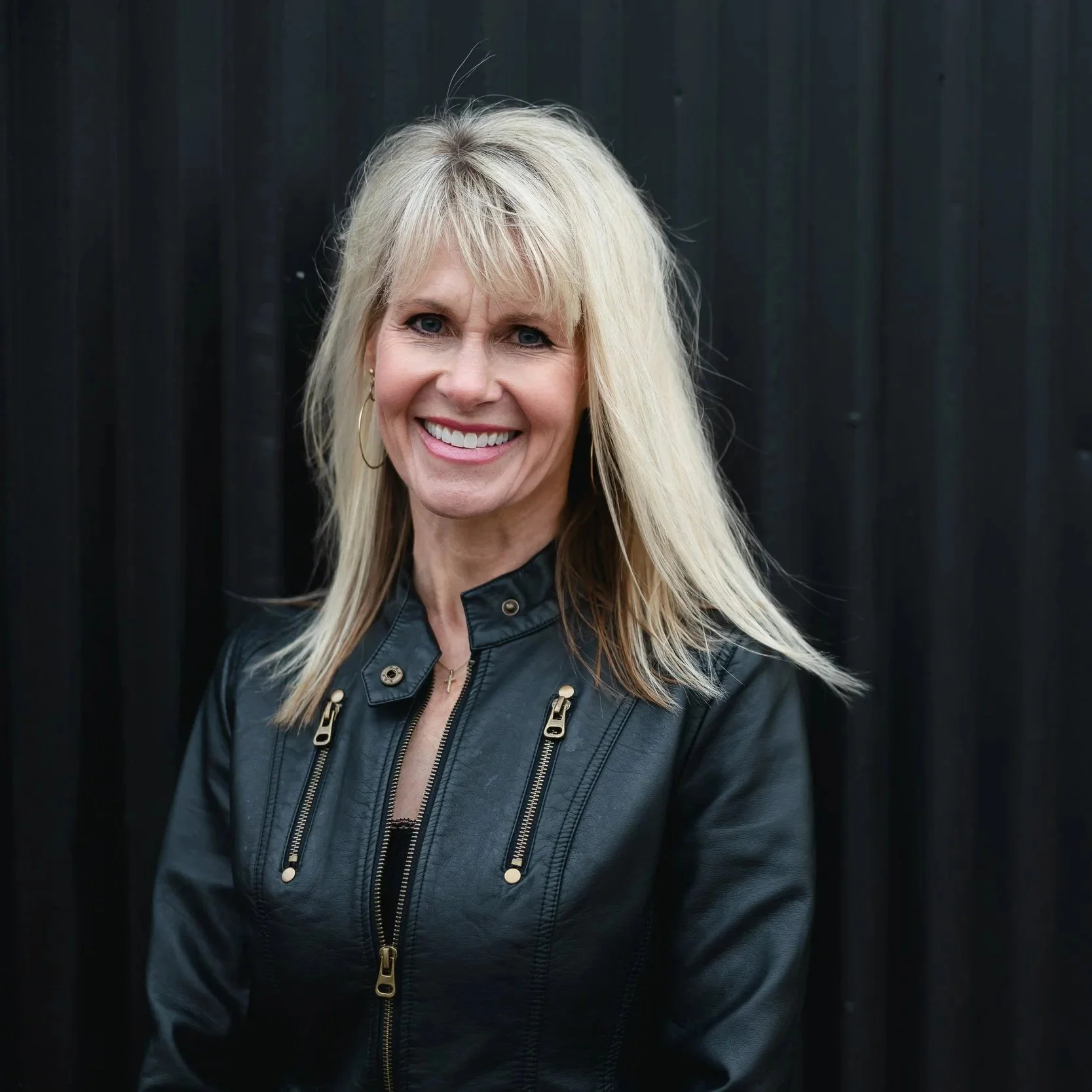 Author Laurie Geisz is smiling and wearing a black leather jacket against a black background.