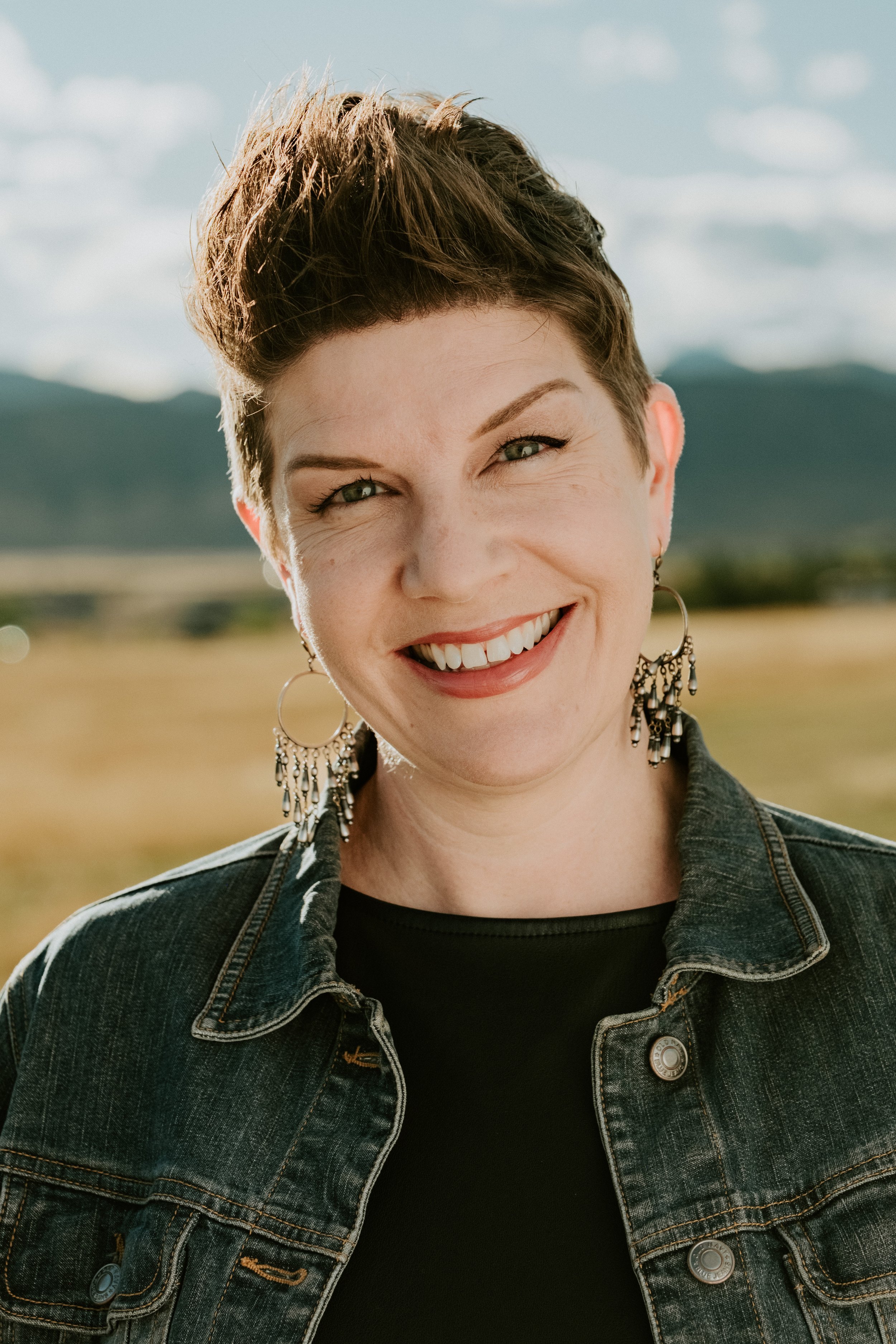 A woman with short brown hair smiling outdoors with mountains and fields in the background.