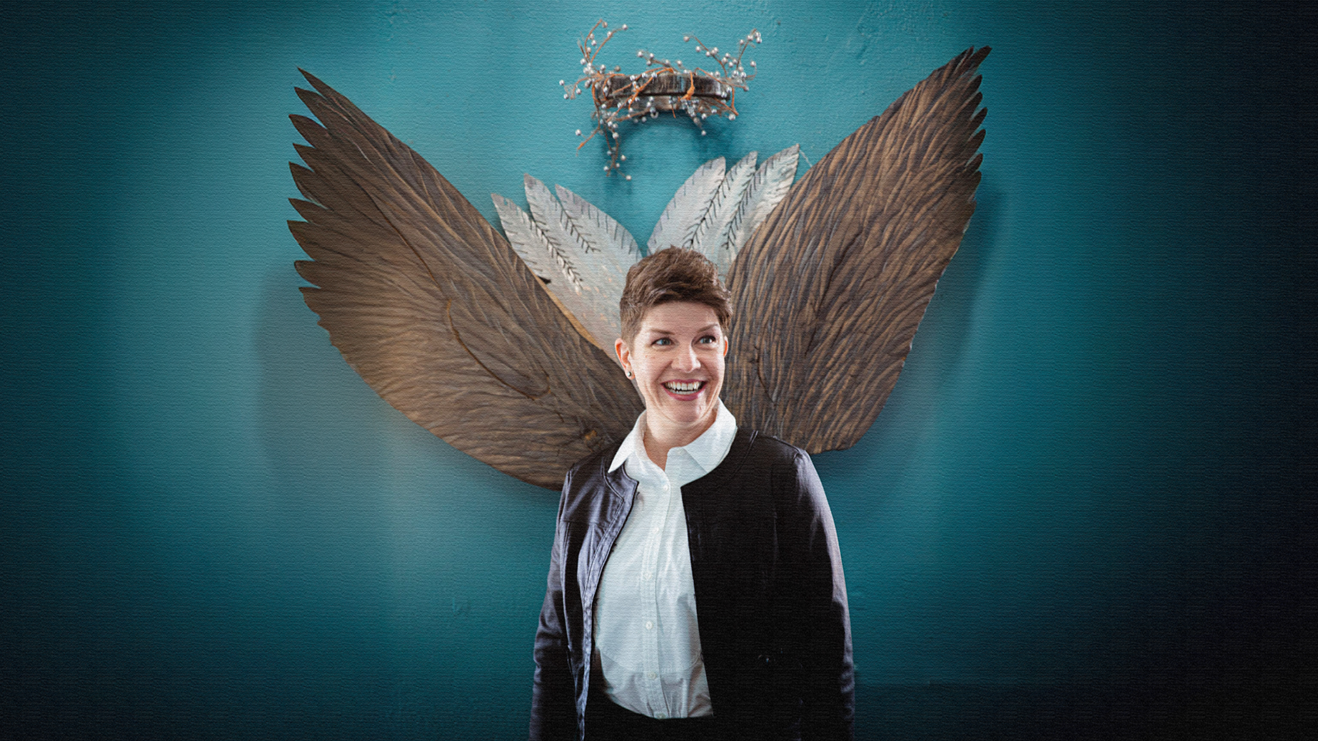 A smiling woman with short brown hair stands in front of a teal wall with a butterfly-shaped art installation made of large brown and white wings, feathers, and a twig crown.