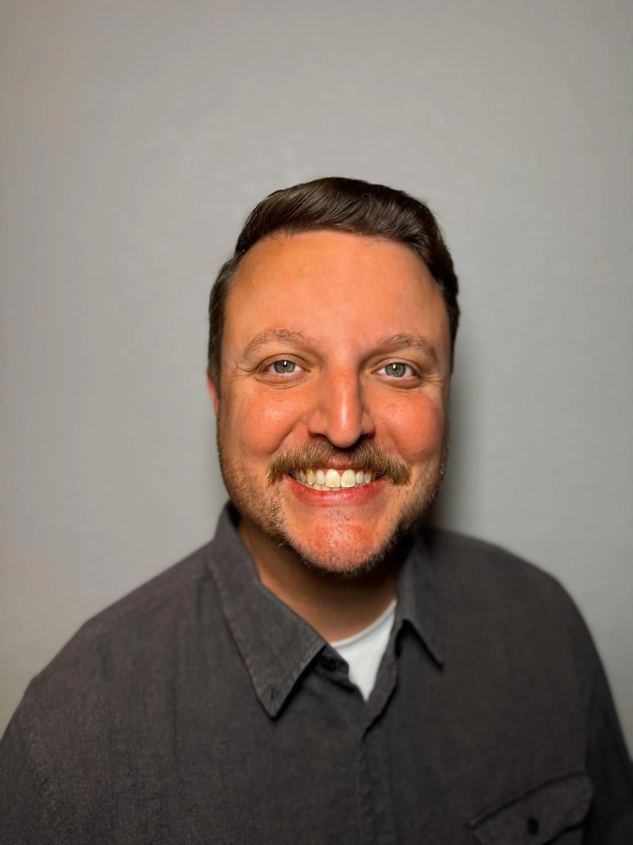 A man is smiling and wearing a dark brown shirt against a gray background.