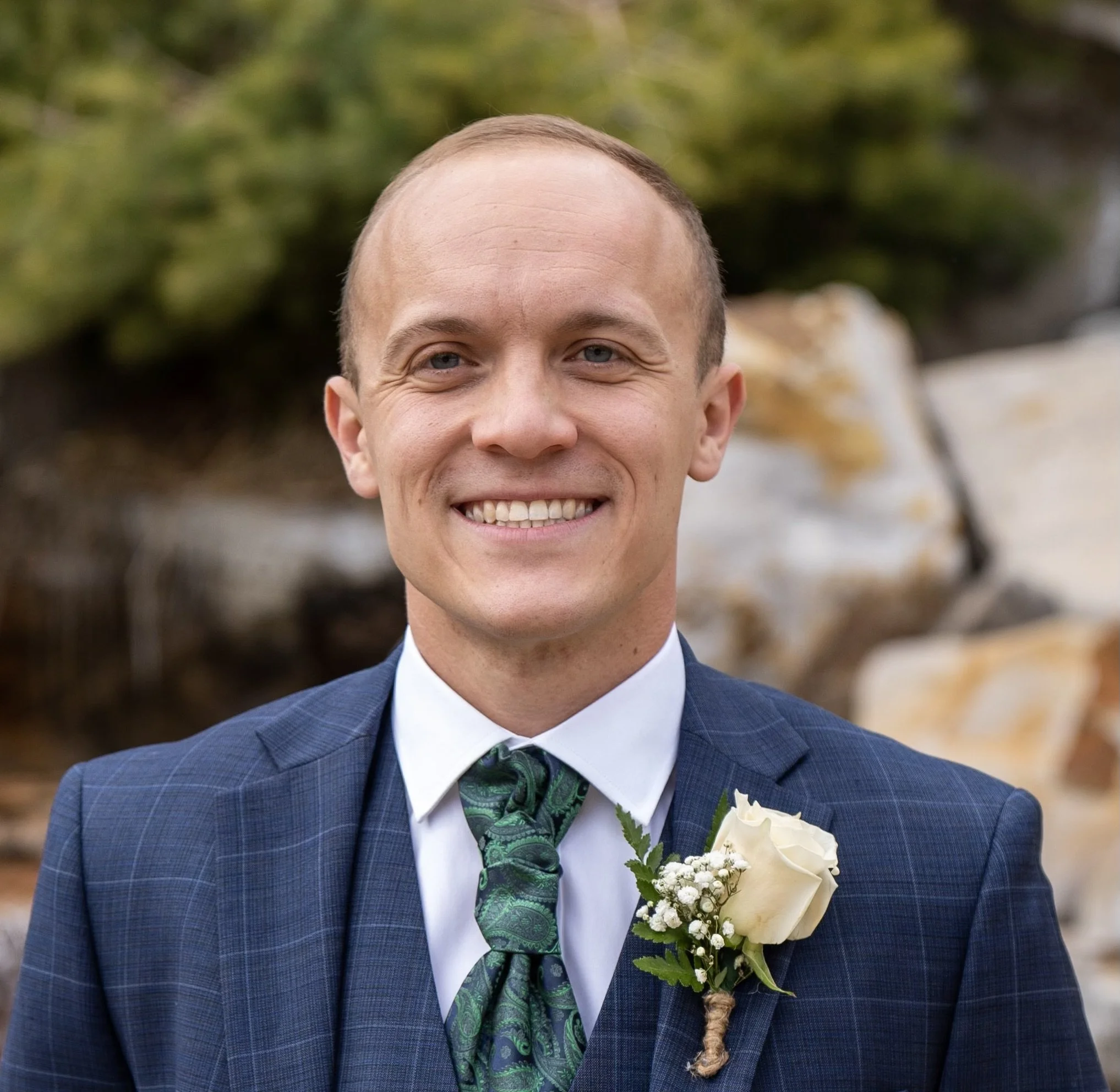 Photo of a man smiling wearing a navy blue suit with a rose in the lapel.