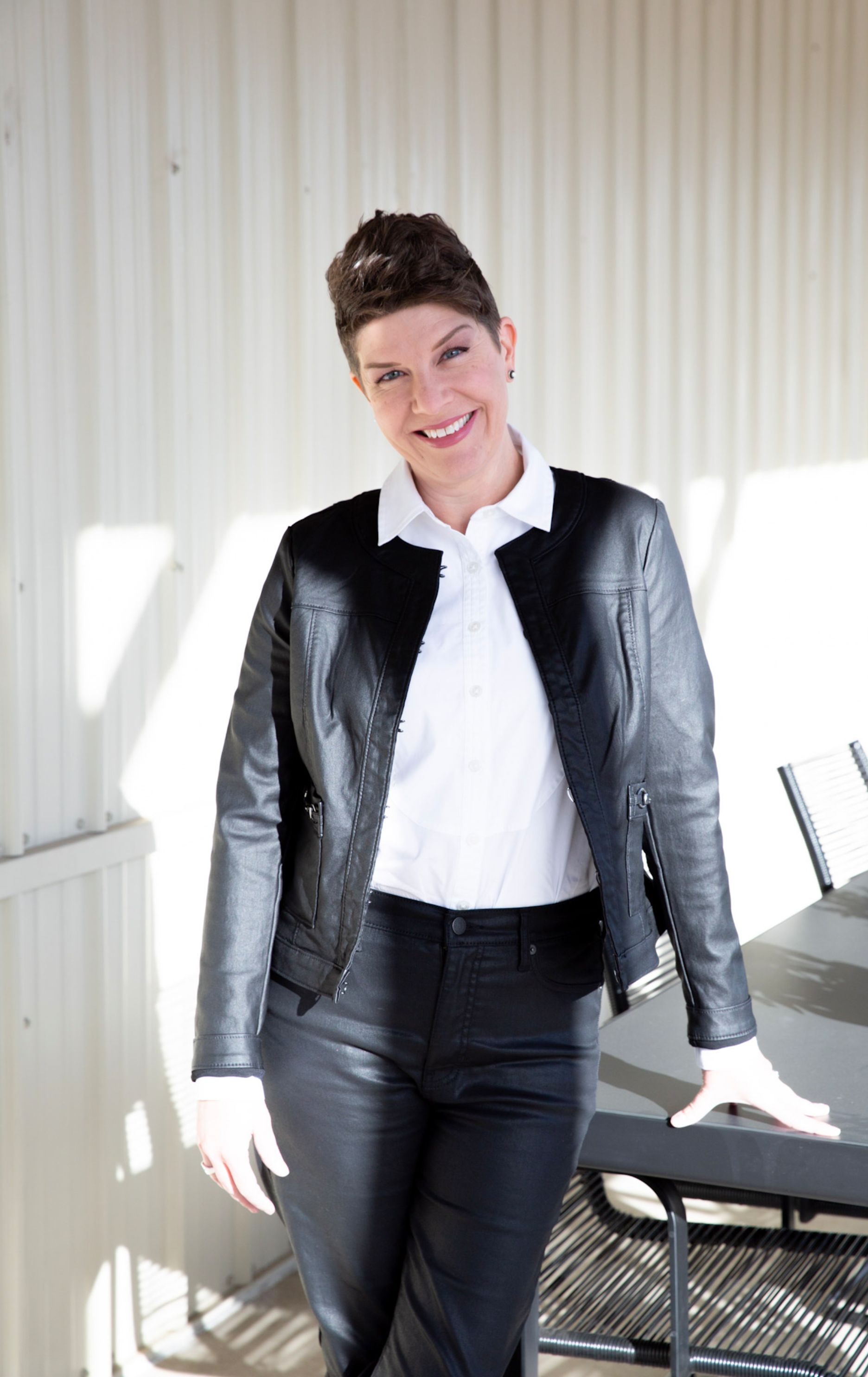 A woman with short brown hair, wearing a black leather jacket, white shirt, and black pants, smiling and standing in a bright room with sunlight coming through the window.