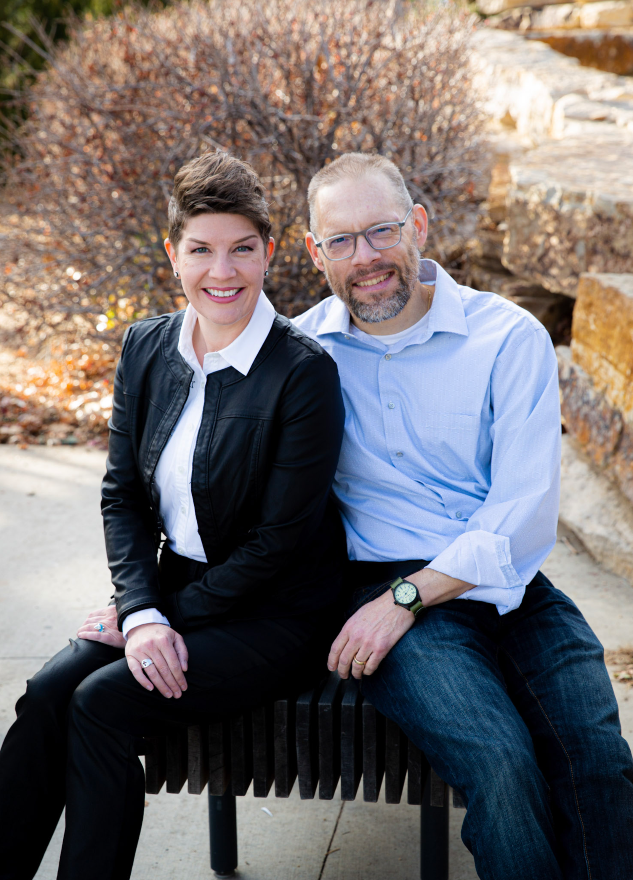 Chaplain Eva Bleeker and her husband Josh are pictured smiling, sitting on a bench outdoors near a bush with brown leaves.