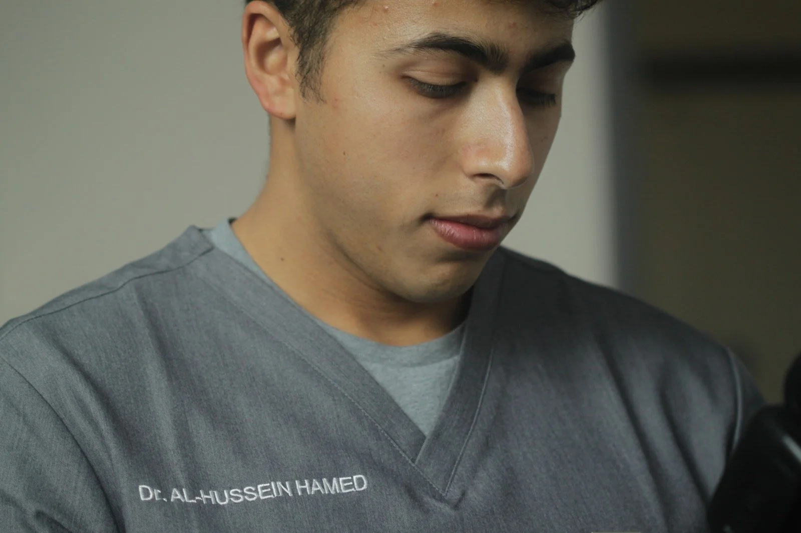 Close-up of a young man wearing a gray medical scrub with the name 'Dr. Al-Hussein Hamed' embroidered on it. He appears to be looking down, possibly at a medical instrument or device.