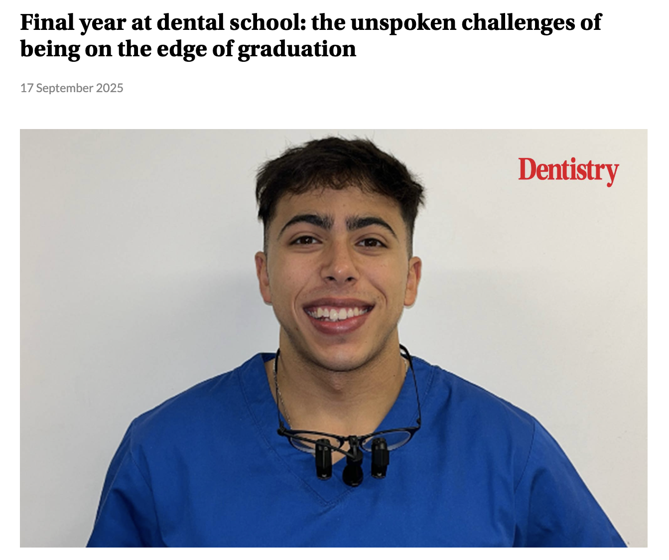 A young male dentist in blue scrubs smiling, with dental loupes hanging around his neck, standing against a plain white wall.