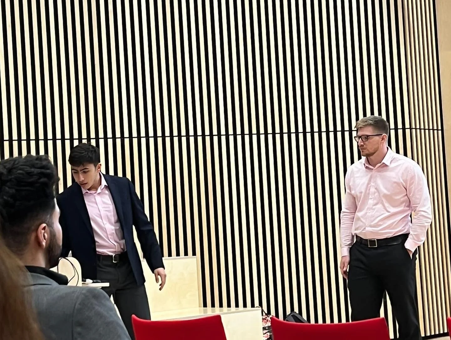 Three men in business attire in a room with a striped black and beige wall, engaged in conversation or presentation, with red chairs and a beige podium visible.