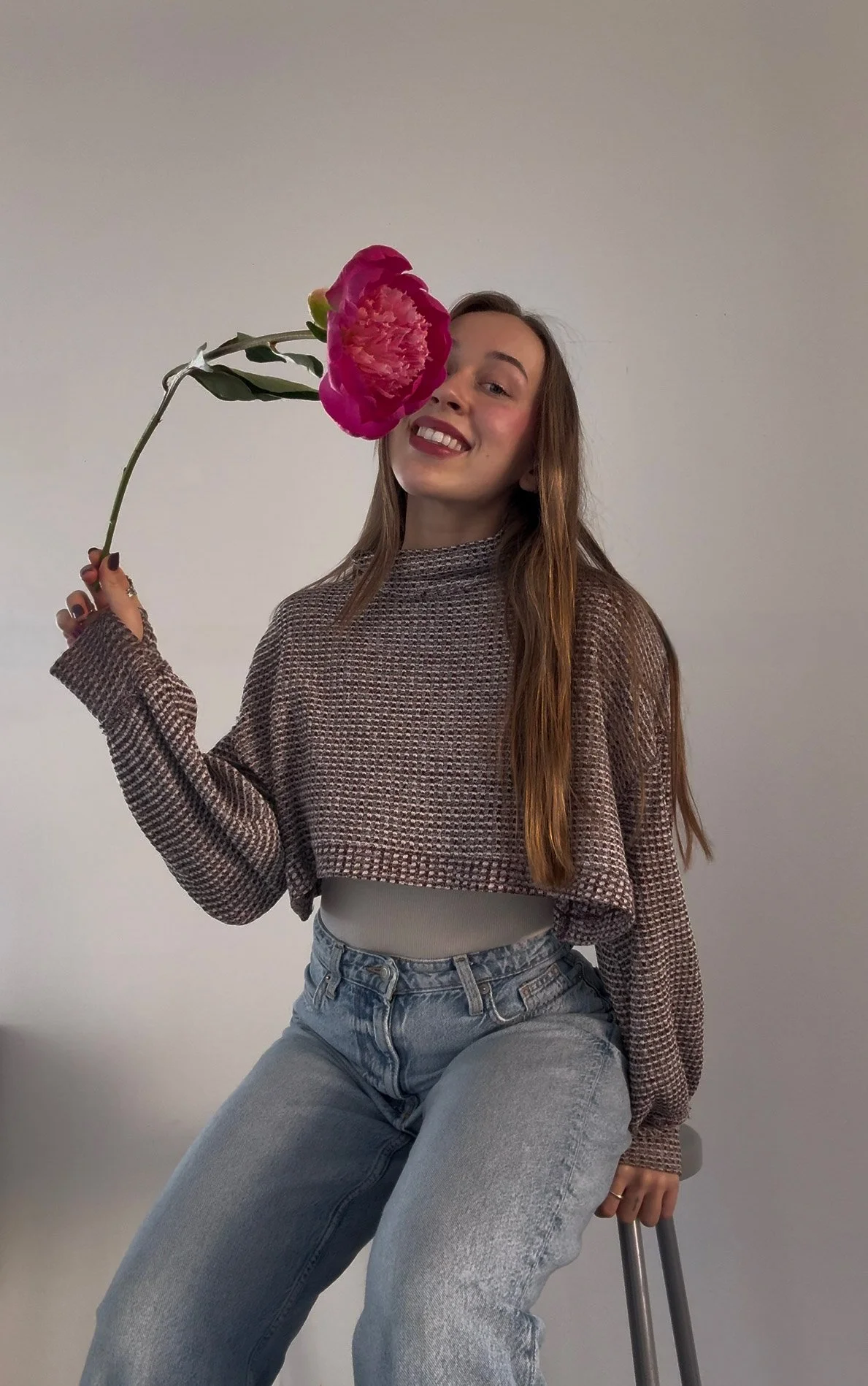 A young woman with long brown hair, wearing a patterned sweater and jeans, sitting on a stool, holding a large pink peony near her face, smiling against a plain light-colored background.