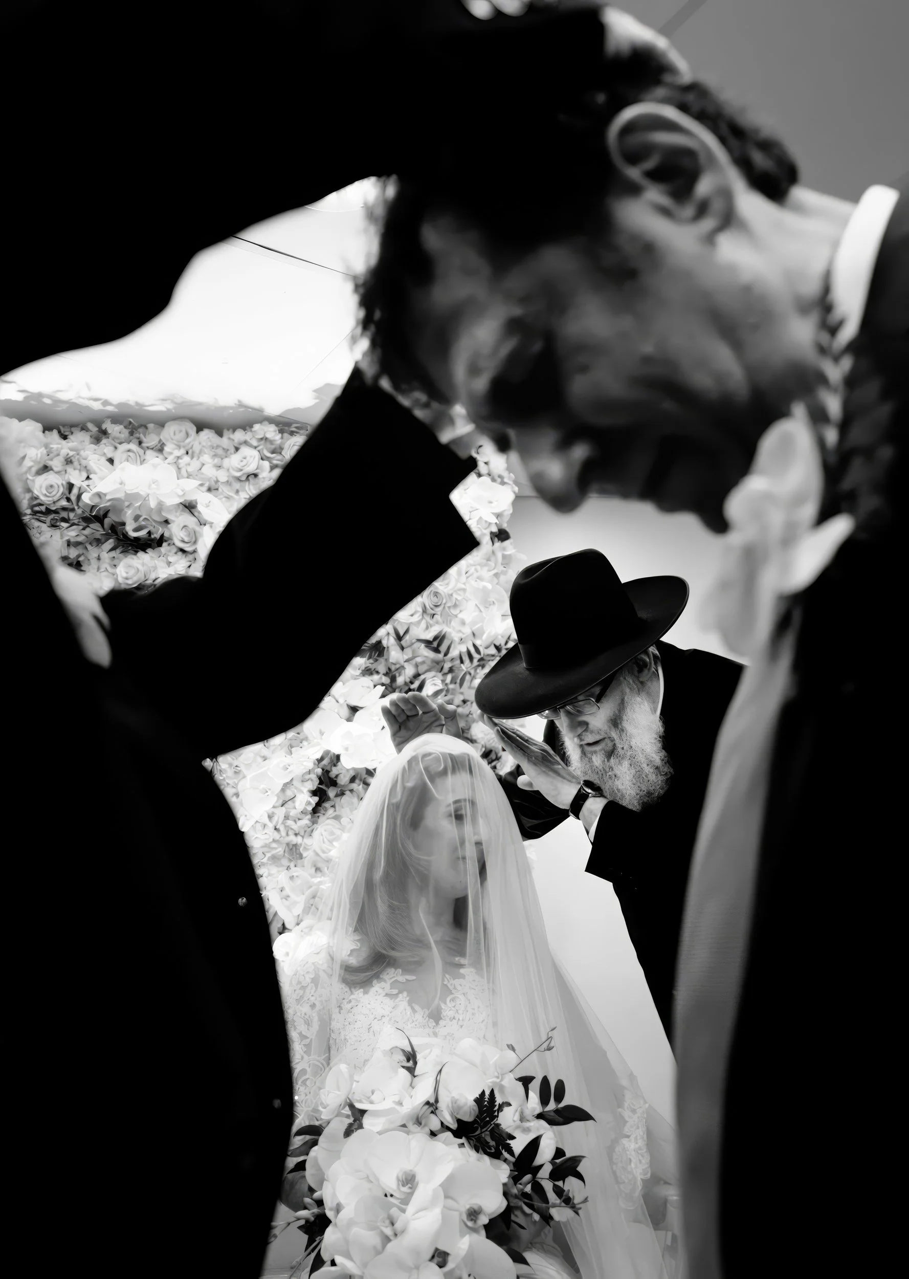 A black and white photo of a wedding ceremony, showing a bride with a veil holding a bouquet of flowers, surrounded by several men, including a rabbi leading the ceremony, all in traditional attire.