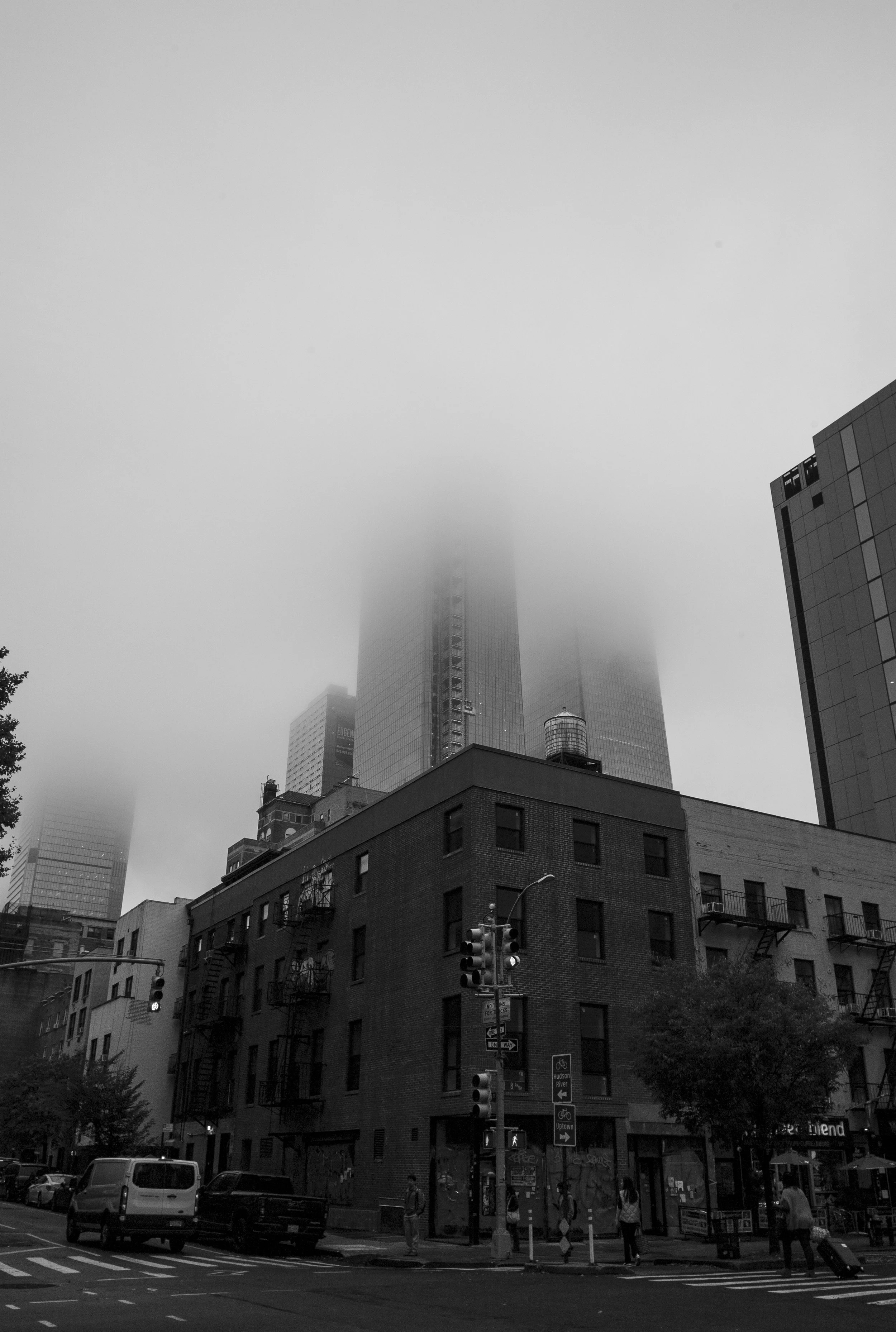 Black and white photo of a city street corner with pedestrians, vehicles, and traffic lights. Tall buildings in the background are obscured by fog or low clouds.