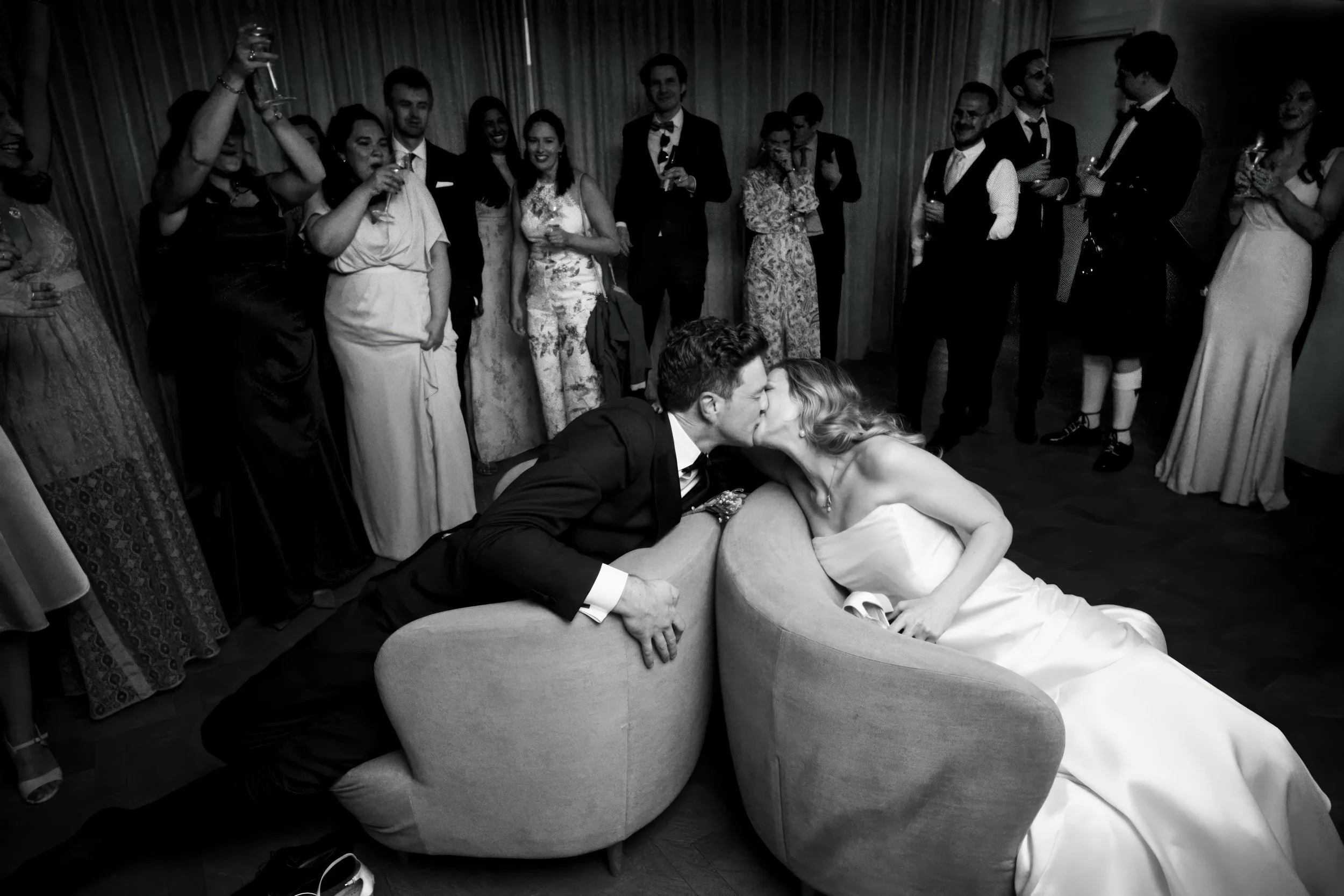 A black and white photo of a wedding reception with a bride and groom sharing a kiss while sitting on a curved couch. Guests in formal attire stand in the background watching and celebrating.