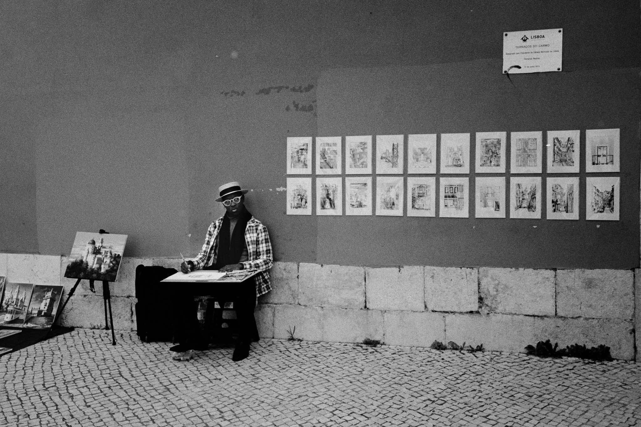 Street artist sitting at a small table on a cobblestone sidewalk, drawing or painting, with artwork displayed on a wall behind him. There are paintings on the ground beside him and a small sign on the wall indicating Lisbon.