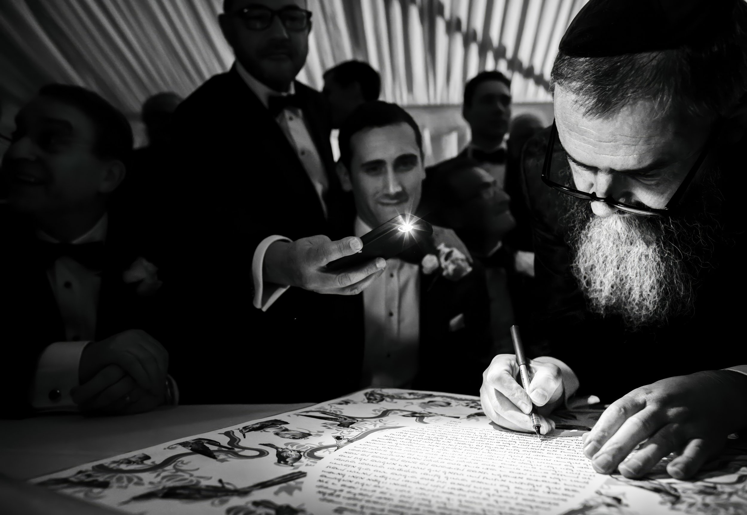 A man with glasses and a long beard signs a document while a group of men in tuxedos, some with bow ties, gather around watching. One man is holding a phone taking a picture or video, with the screen shining. The setting appears to be formal, possibl