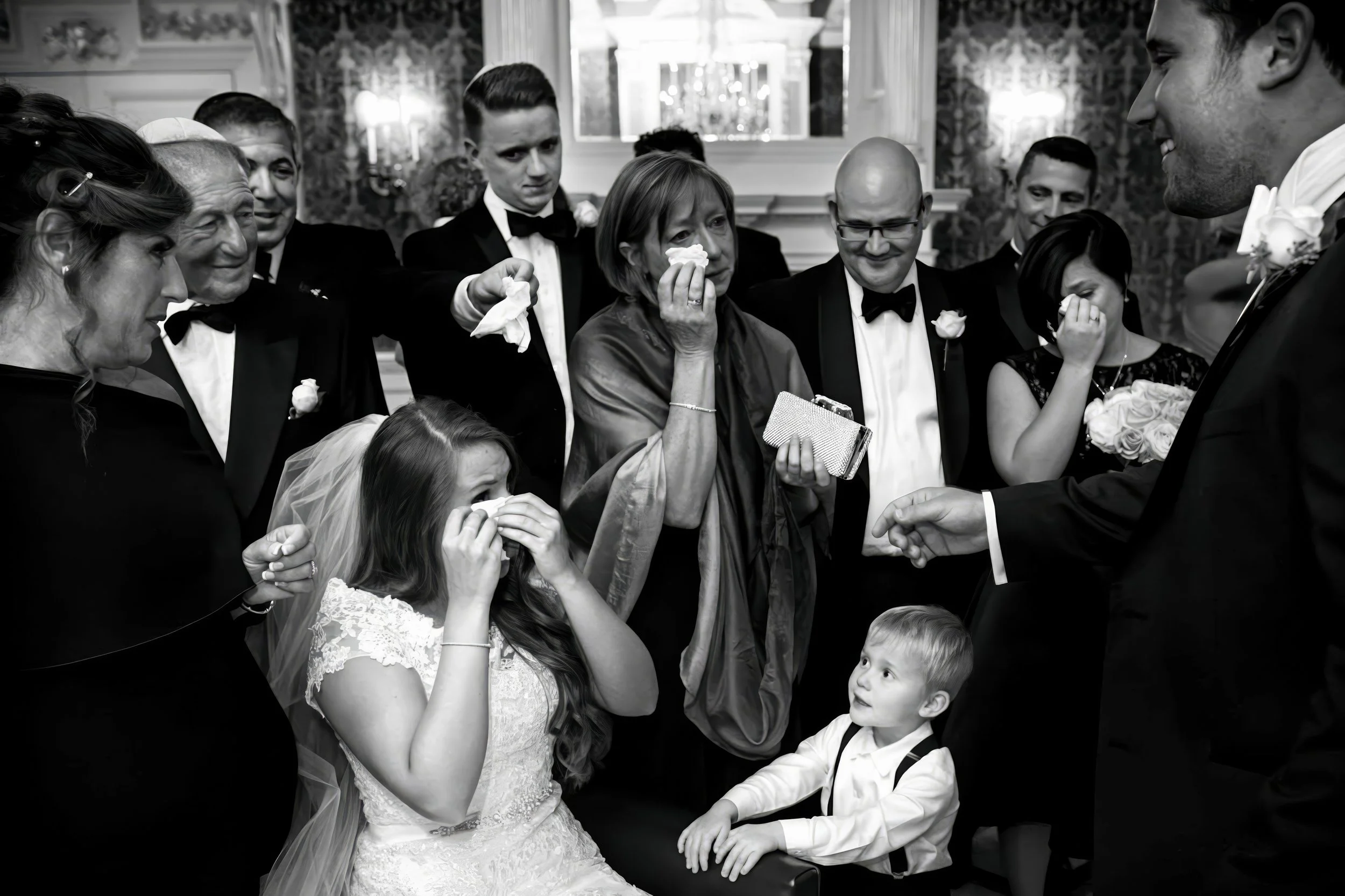 Black and white photo of a wedding scene with crying and emotional guests surrounding a bride and groom. The bride, seated and dressed in a wedding gown, is holding her face and crying, while a young boy sits in front of her looking at her. The guest