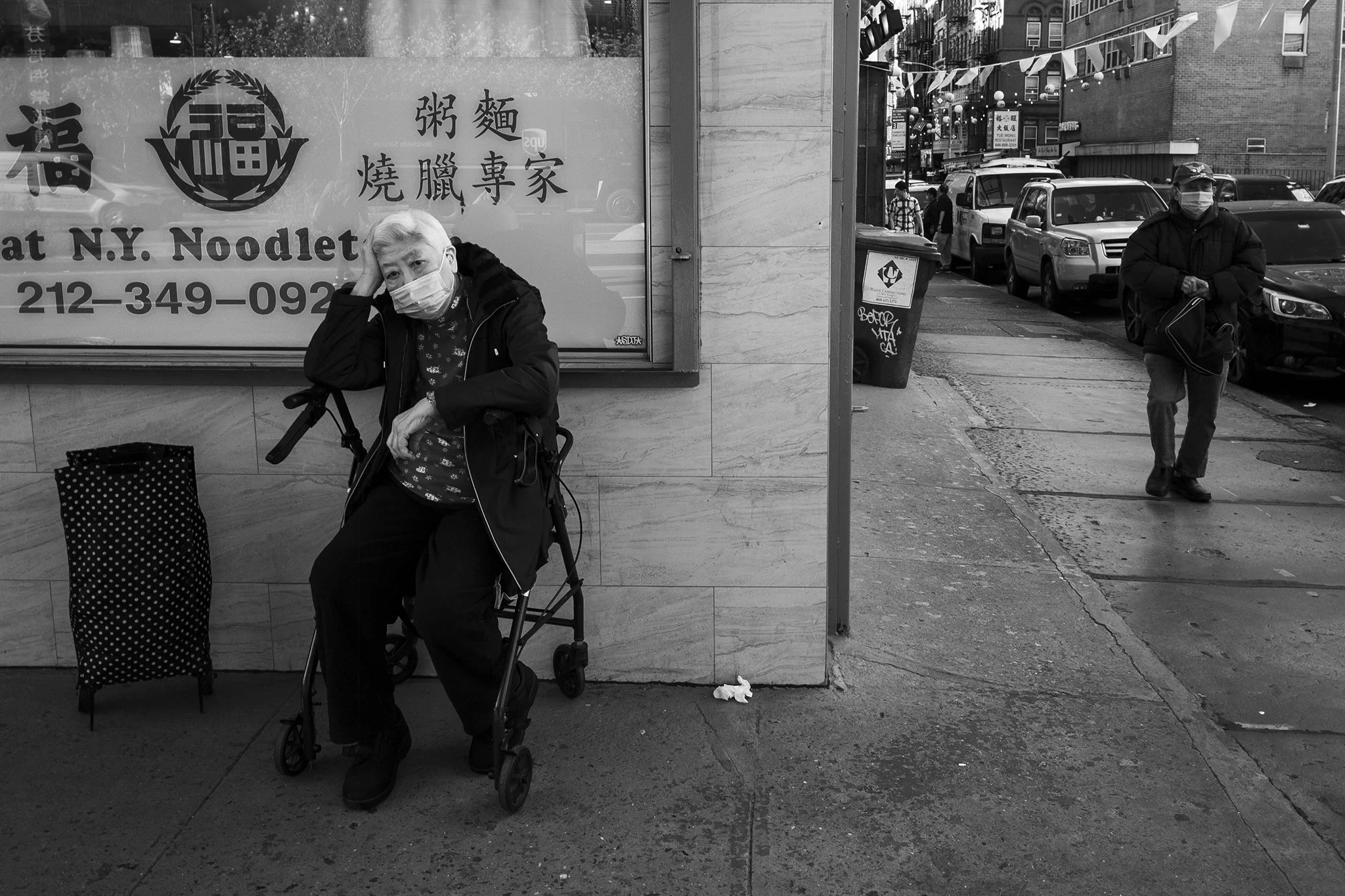 An elderly woman with a walker and face mask sitting on a sidewalk, with a shopping bag beside her. A man with a face mask and bag walking on the sidewalk in an urban street, with parked cars and buildings in the background.