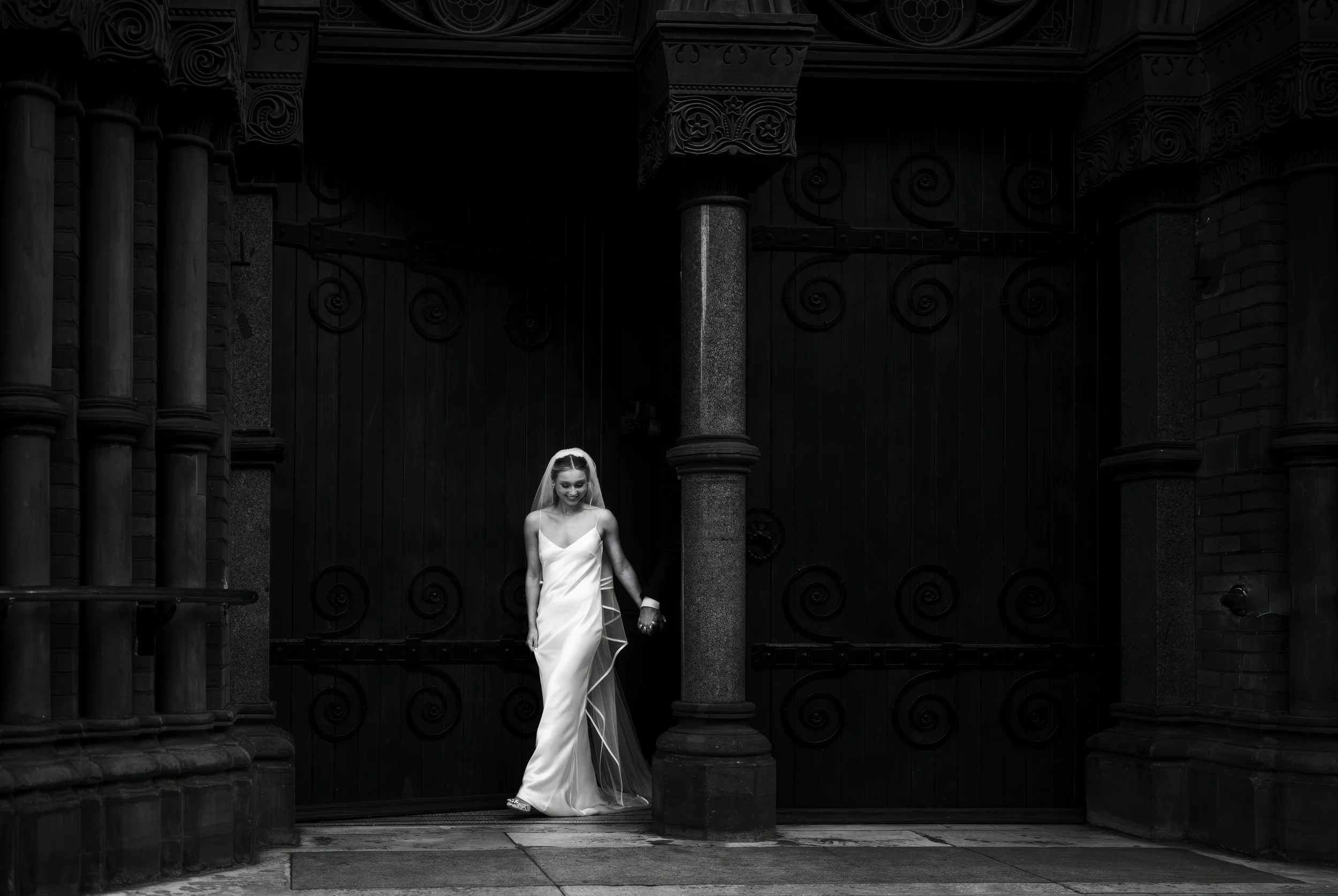 A bride in a white dress and veil standing at a black iron gate, holding hands with someone outside the frame, in front of a dark, ornate building.