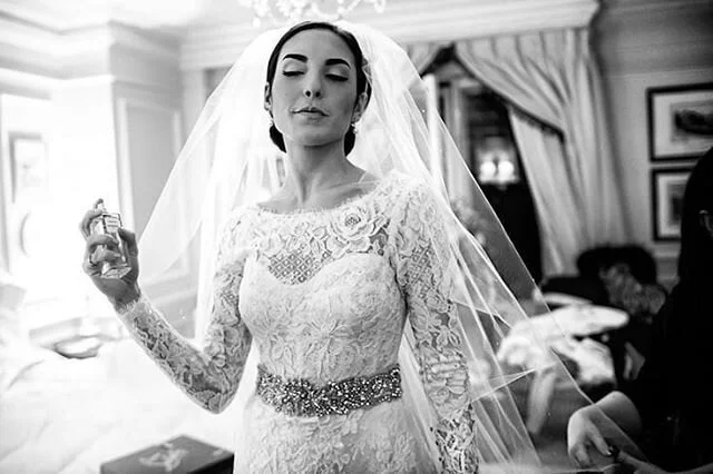 A bride in a lace wedding dress and veil holding a perfume bottle, standing in a room with curtains and framed pictures.