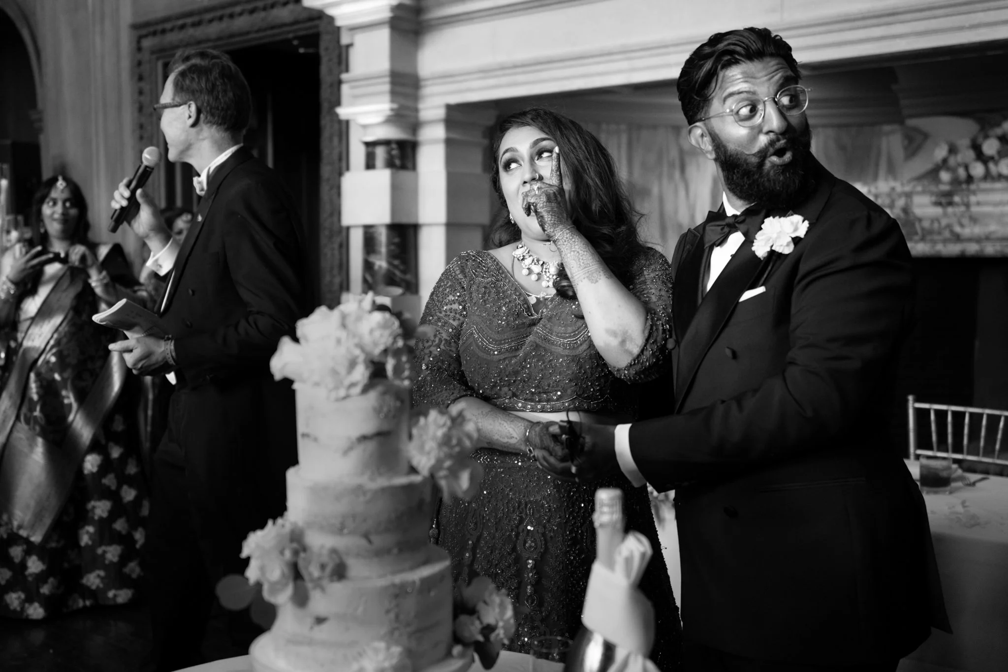Black and white photo of a wedding celebration with a bride and groom. The bride is wearing a sparkling dress and jewelry, with henna on her hand, and is covering her mouth with her hand. The groom is dressed in a tuxedo with a flower on his lapel, making an expressive face. In the background, a man in glasses and a tuxedo is speaking into a microphone, and a woman is playing a guitar. A wedding cake with flowers is in the foreground.