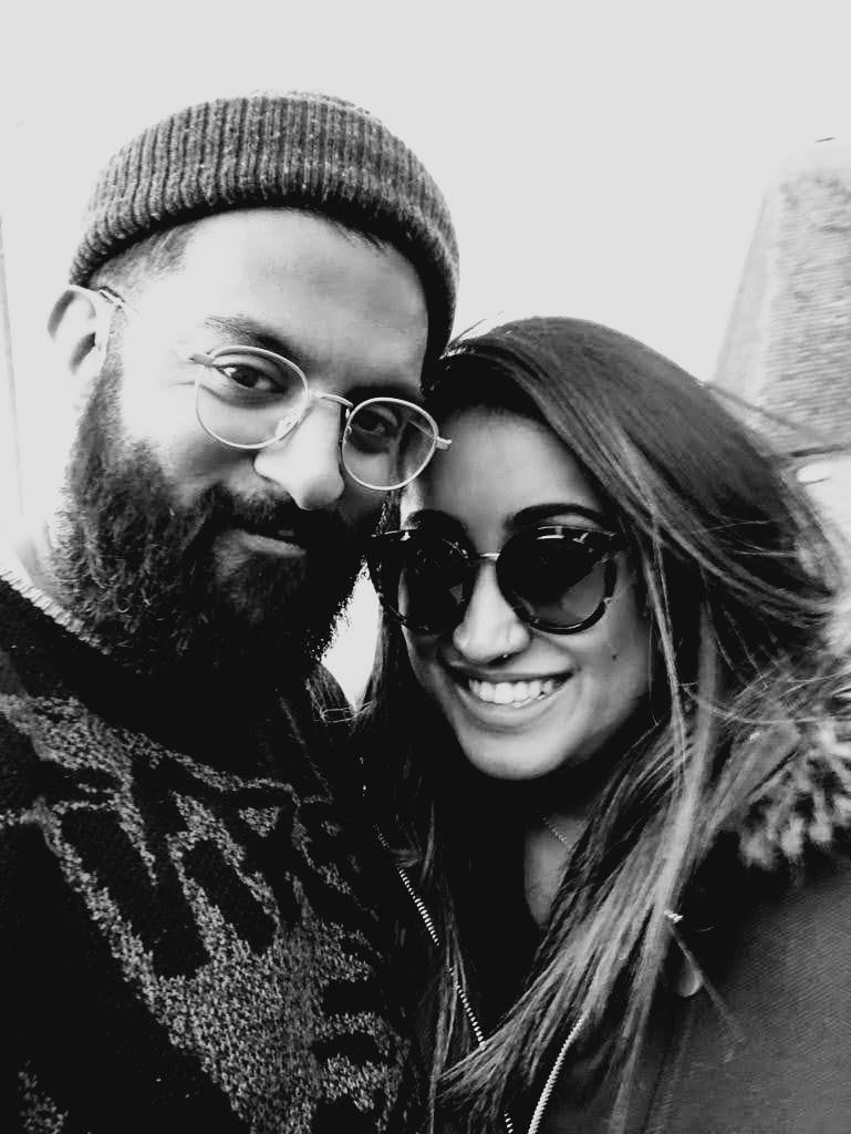 A black and white photo of a smiling man and woman taking a close selfie outdoors. The man has a beard, glasses, and is wearing a knit beanie, while the woman has long hair and is wearing sunglasses.