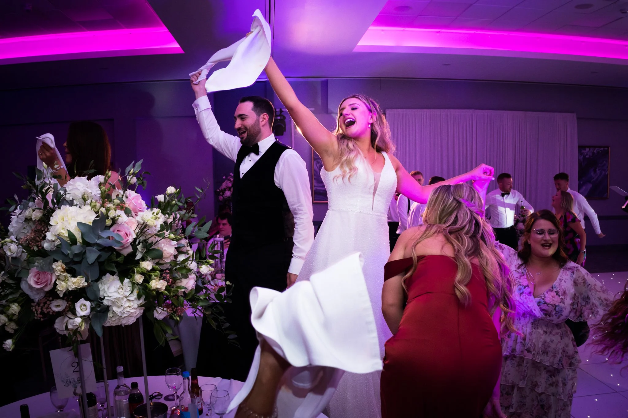 Bride and groom dancing at wedding reception with friends, floral arrangements, and purple lighting