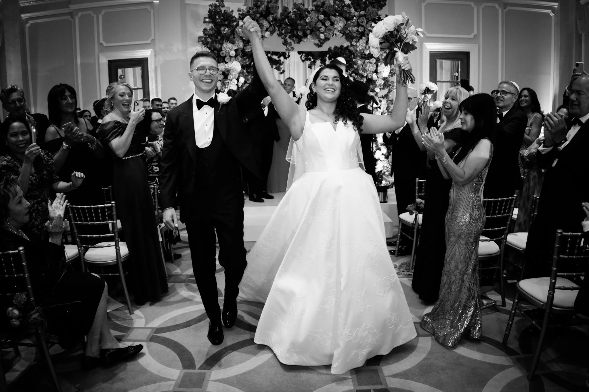 A newlywed couple walking down the aisle at their wedding reception, surrounded by cheering guests, with the bride holding a bouquet and the groom wearing a tuxedo.
