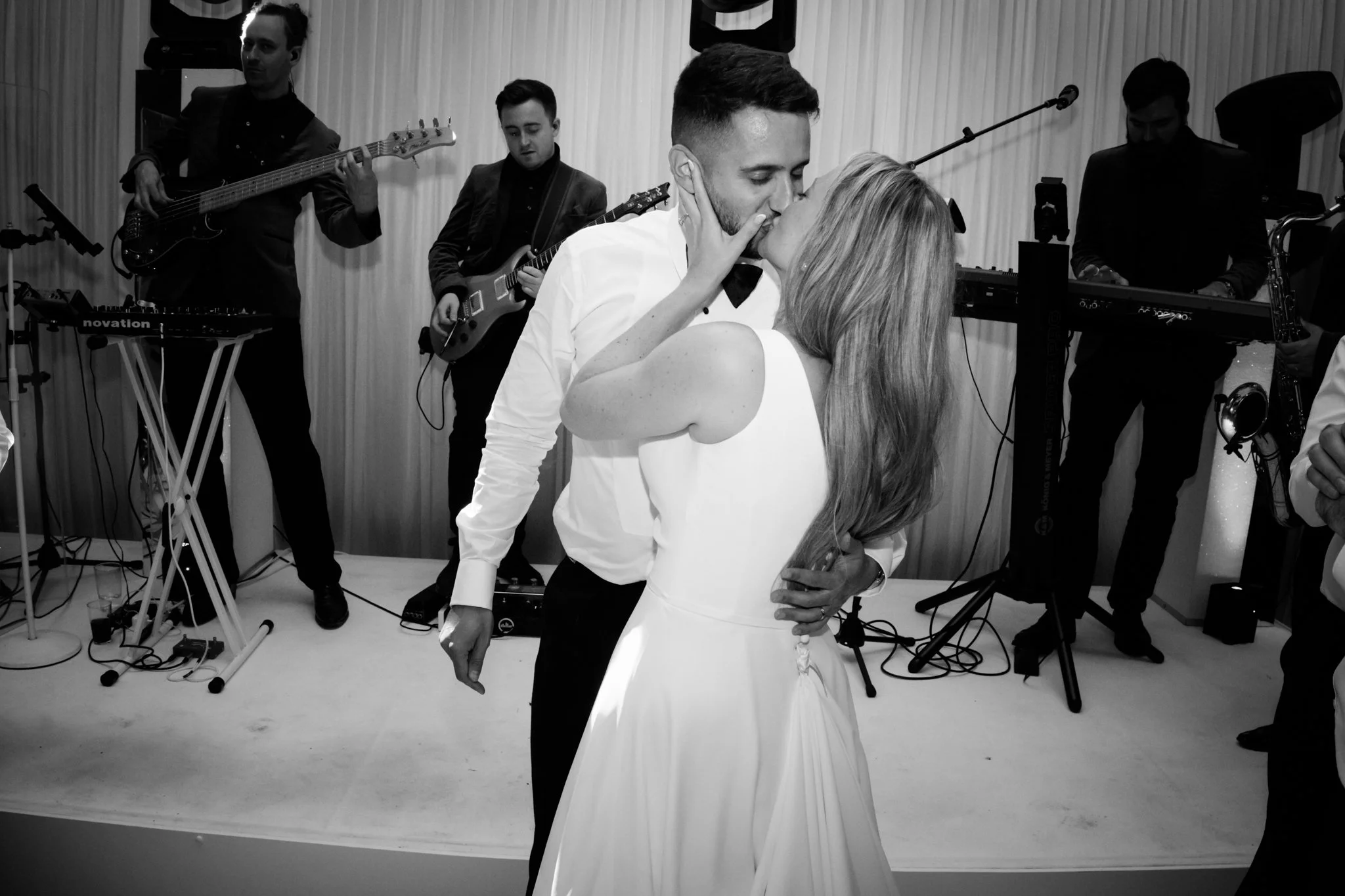 A black and white photo of a newlywed couple kissing during their wedding reception, with a live band playing in the background.
