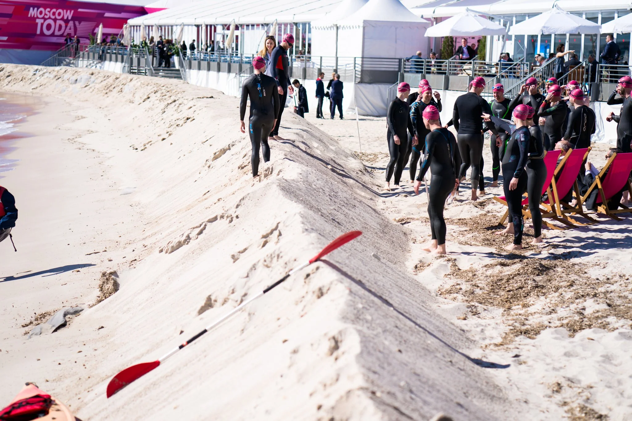 Swimmers in black wetsuits with pink swim caps gather on a sandy beach before a race, with a pink banner reading 'Moscow Today' in the background.