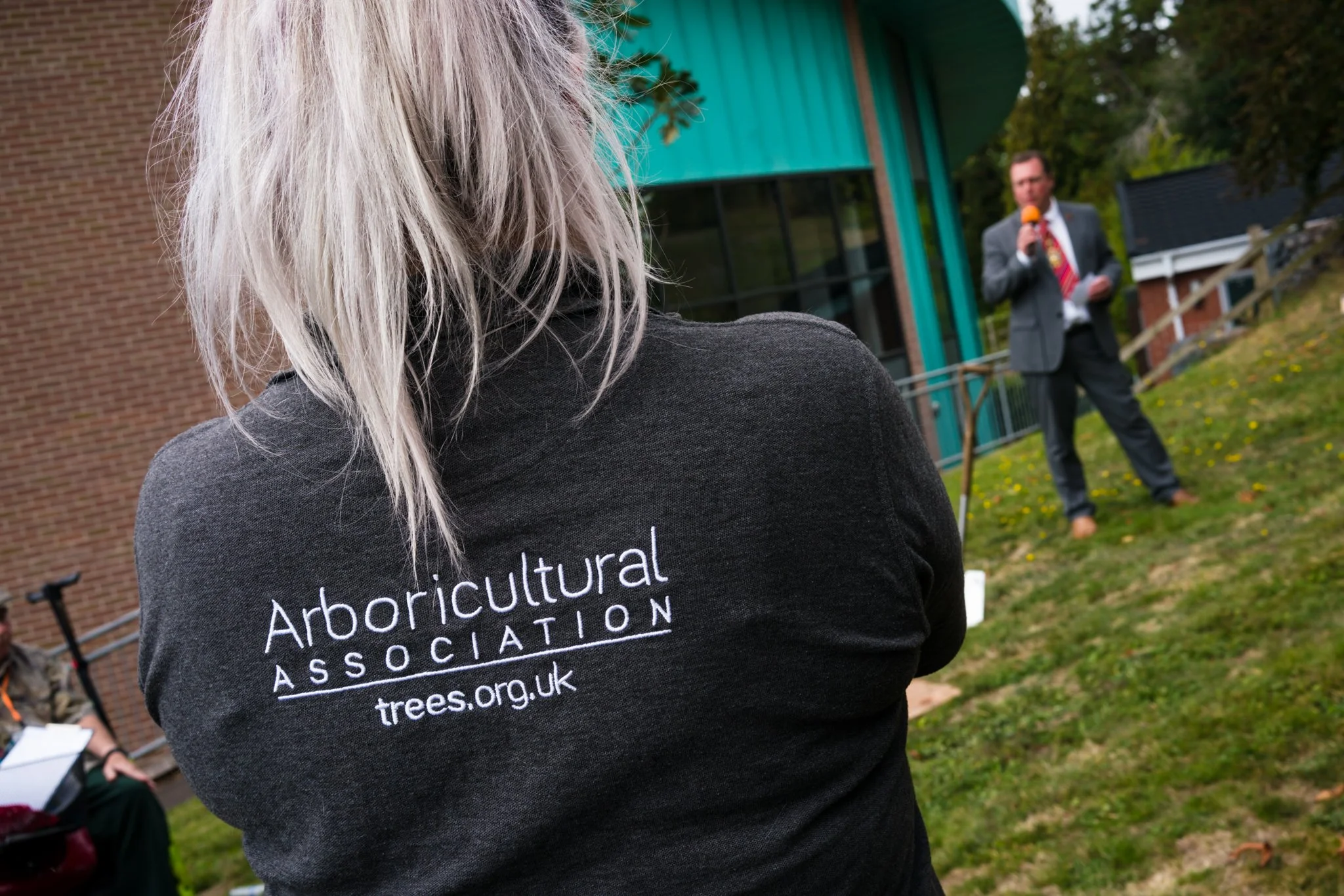 Person wearing a black hoodie with 'Arboricultural Association' logo on the back, standing outdoors at a gathering, with a man in a suit speaking into a microphone in the background
