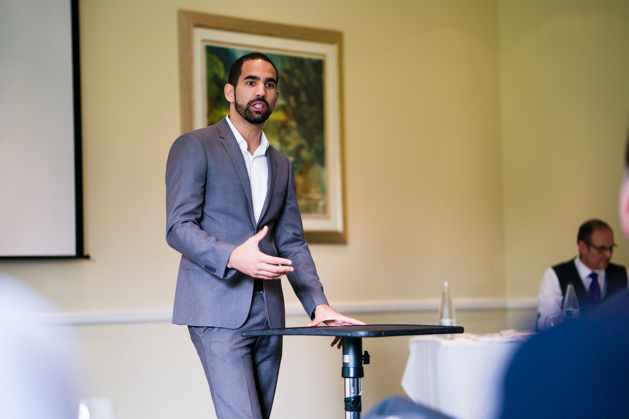 A man in a gray suit giving a presentation in a conference room, with a man sitting at a table in the background.