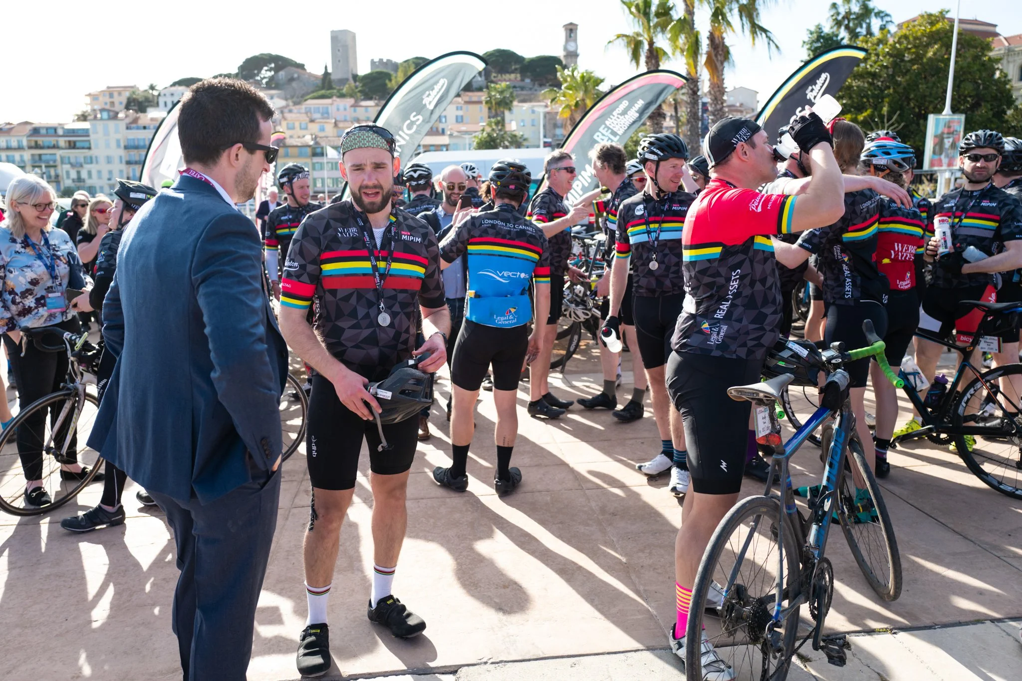 Group of cyclists wearing black jerseys with rainbow stripes, some with medals, some drinking water, gathered outdoors with bicycles and flags in the background, on a sunny day.
