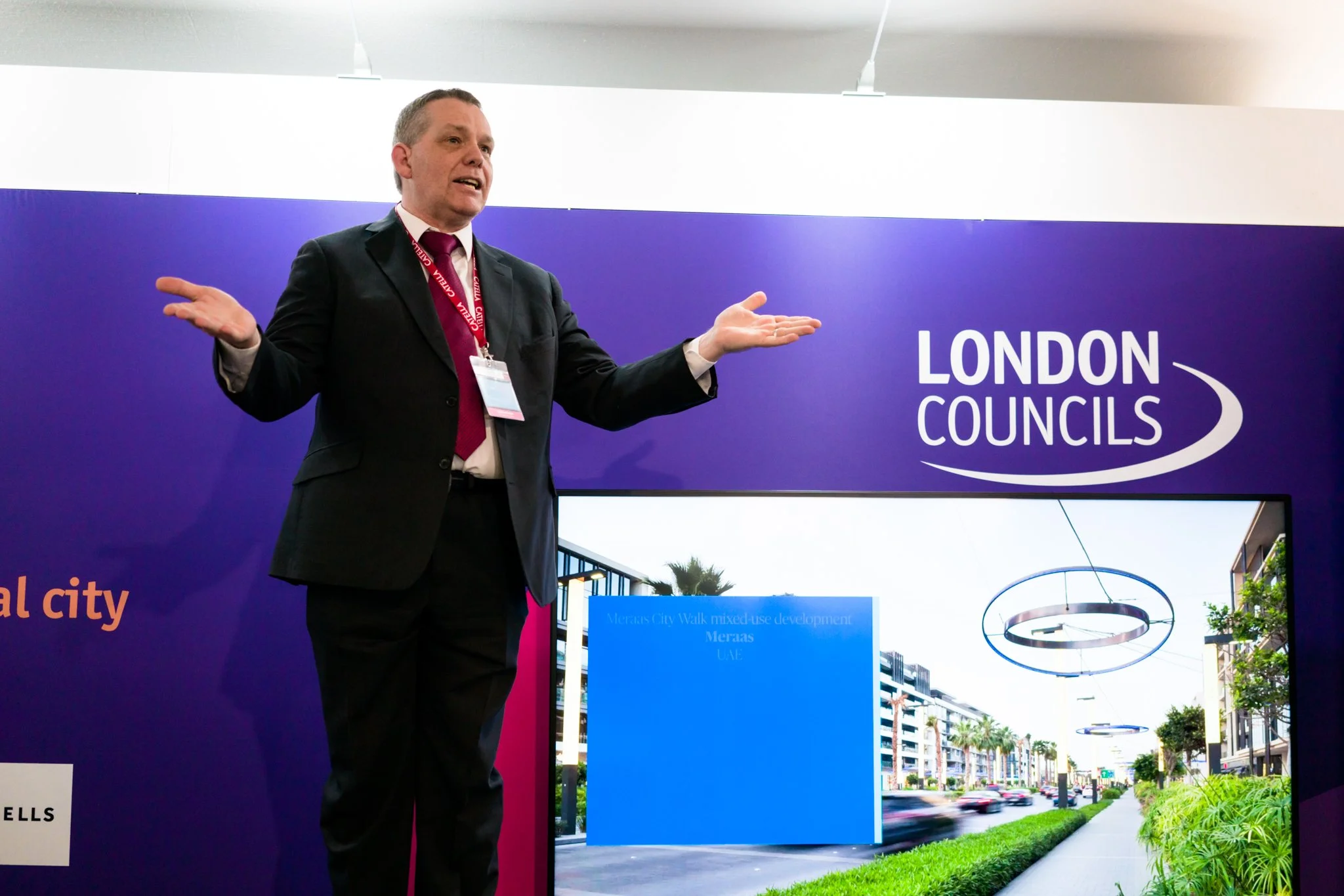 A man in a black suit with a red tie and a conference badge is speaking with his arms open at a London Councils event, with a purple background and a picture of a modern street scene on a display in front of him.